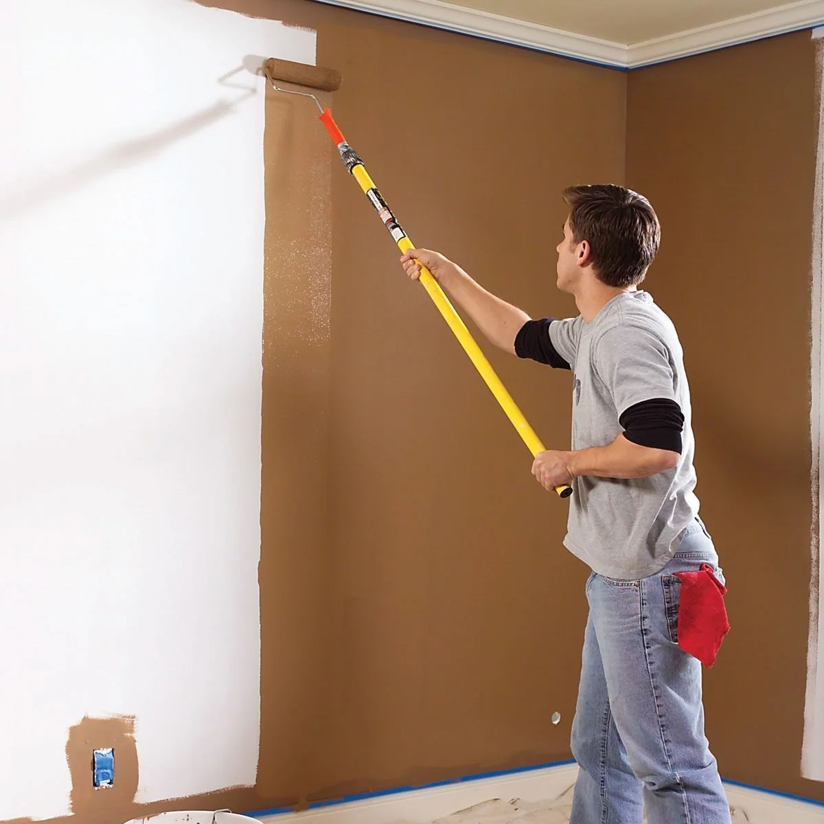 A man painting a wall with a roller brush, applying brown paint over white paint in a room, with a red rag hanging from his pocket.