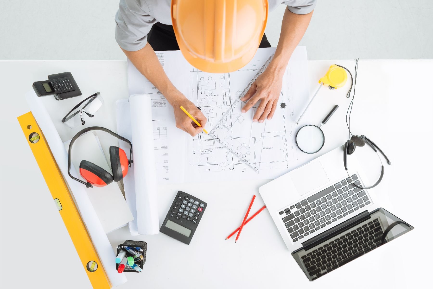 Overhead view of an architect's workspace with blueprints, a yellow spirit level, a calculator, a laptop, a hard hat, a measuring tape, ear protection, and drafting tools, suggesting building design and planning.