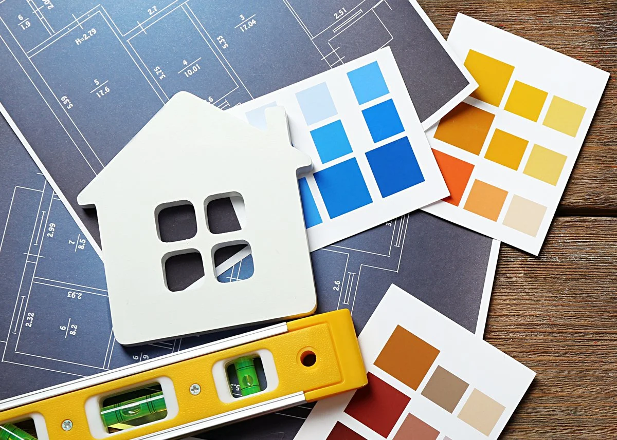 Blueprints, color swatches, a white house cutout, and a yellow level tool on a wooden surface, representing home building or renovation planning.