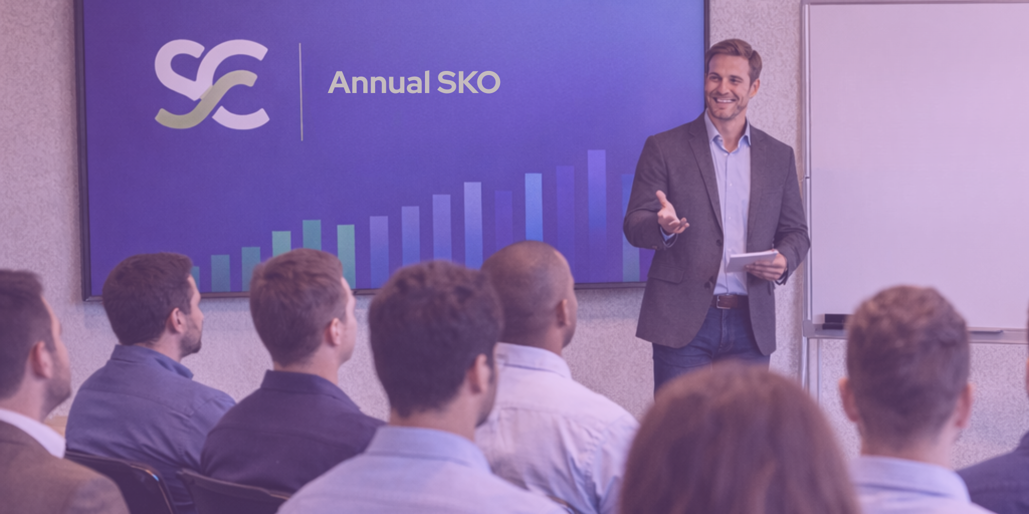 A man in a suit giving a presentation to a group of people in a conference room, with a large screen displaying a logo and the words "Annual SKO" and a bar graph.