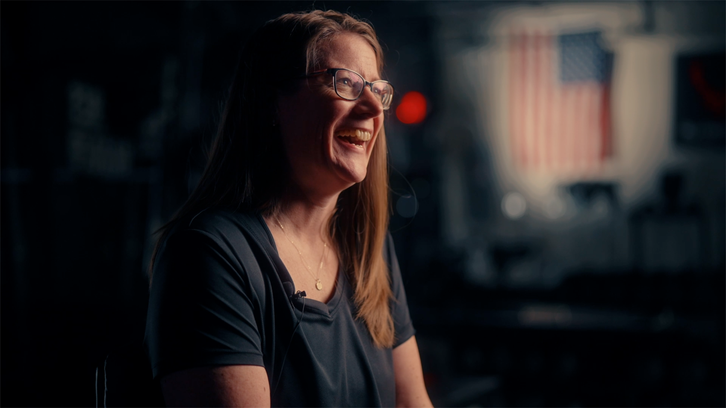 A woman with glasses smiling indoors with windows and blurred lights in the background.