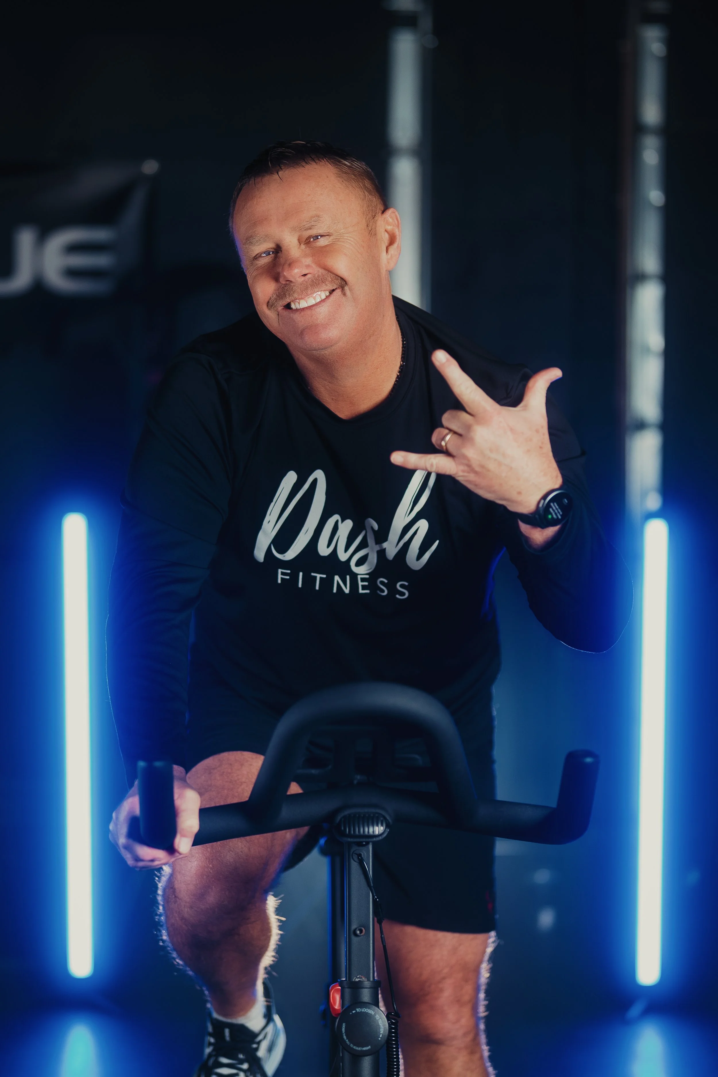 Man in black t-shirt with 'Dash Fitness' logo riding an exercise bike in a dark gym with vertical blue LED lights.