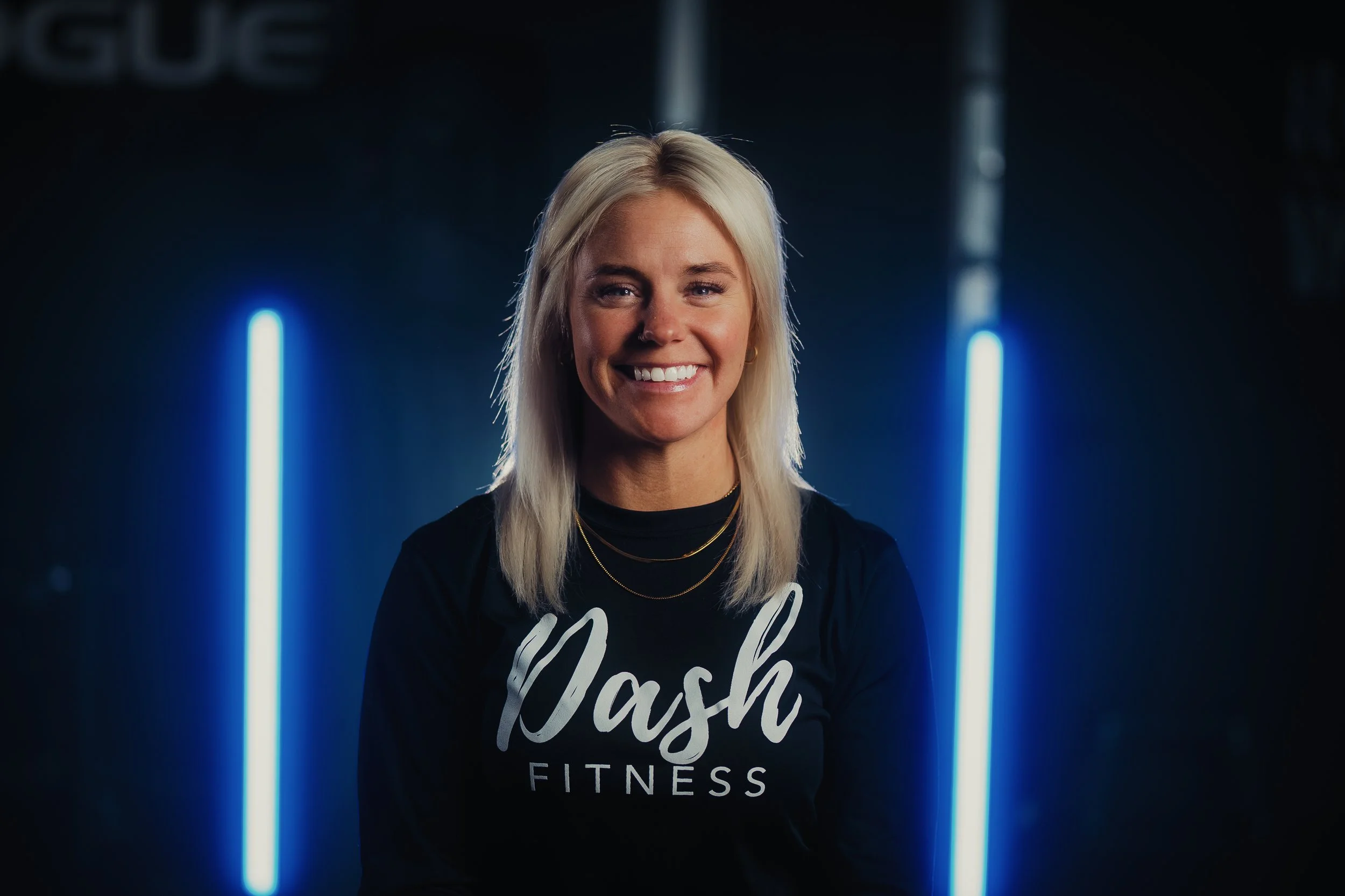 A smiling woman with long blonde hair wearing a black shirt with white text that reads 'Wash Fitness', standing in front of a dark background with two vertical blue neon lights.