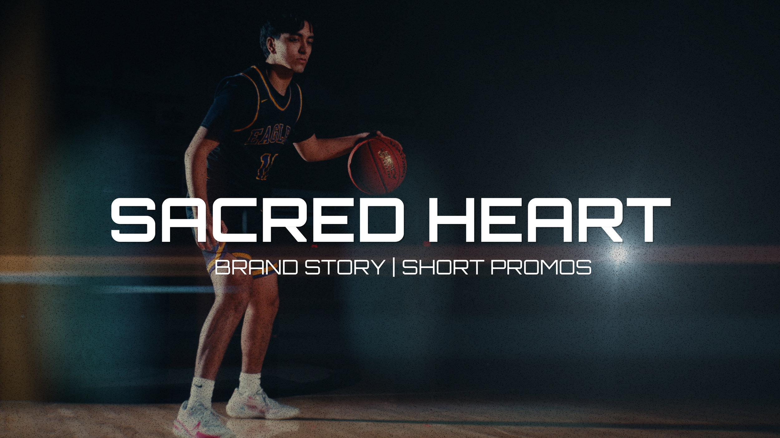 A young male basketball player in a black jersey with yellow accents dribbling a basketball on a court at night, with the words 'Sacred Heart' and 'Brand Story | Short Promos' overlaid on the image.