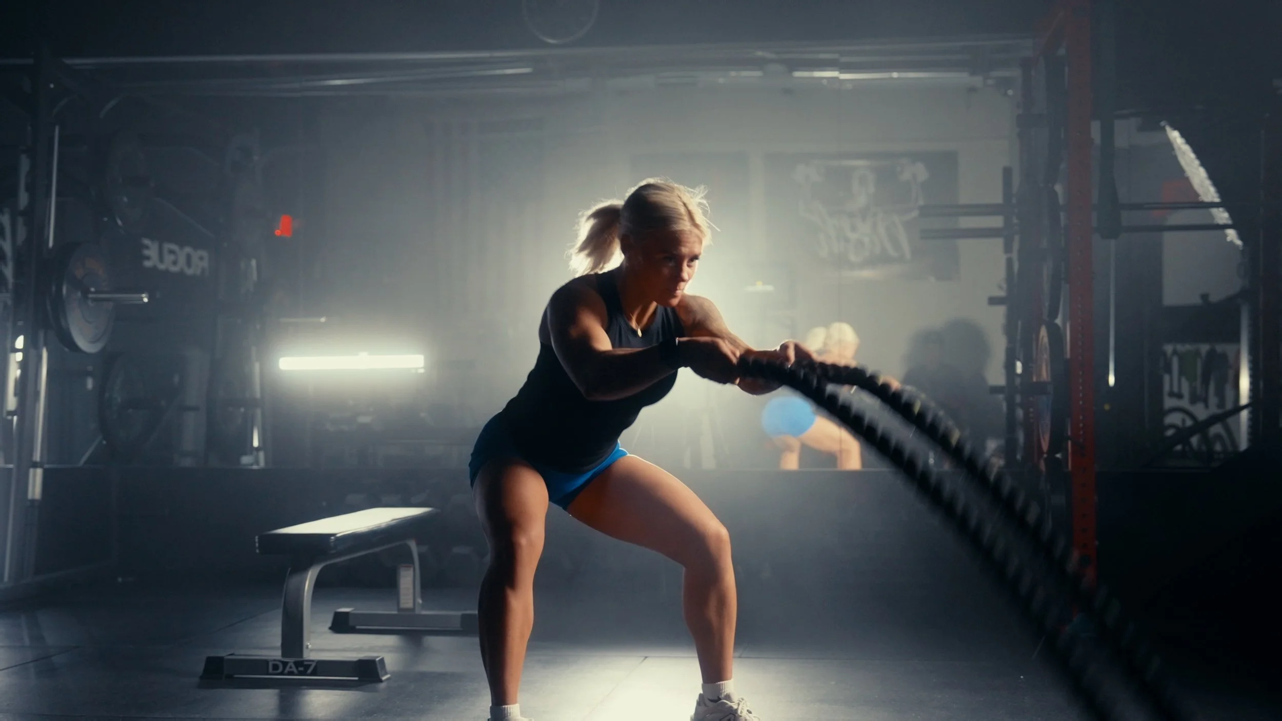 A woman with blonde hair tied in a ponytail wearing a black tank top and blue shorts performing a tire flip exercise in a gym, with gym equipment and weight plates in the background.