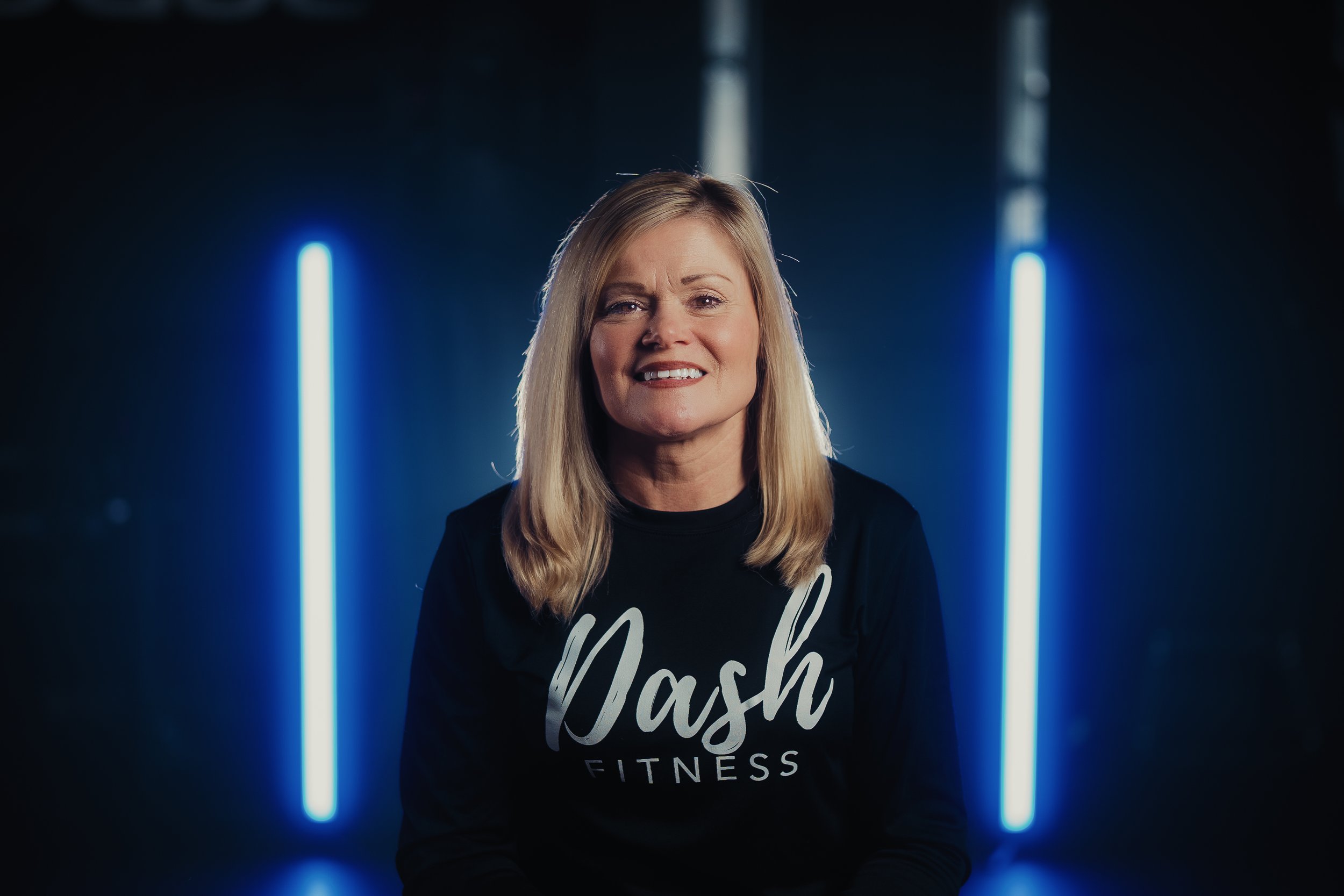 A woman with blonde hair smiling, wearing a black shirt with white text that reads 'Wash Fitness,' standing in front of a dark background illuminated by two vertical neon blue lights.