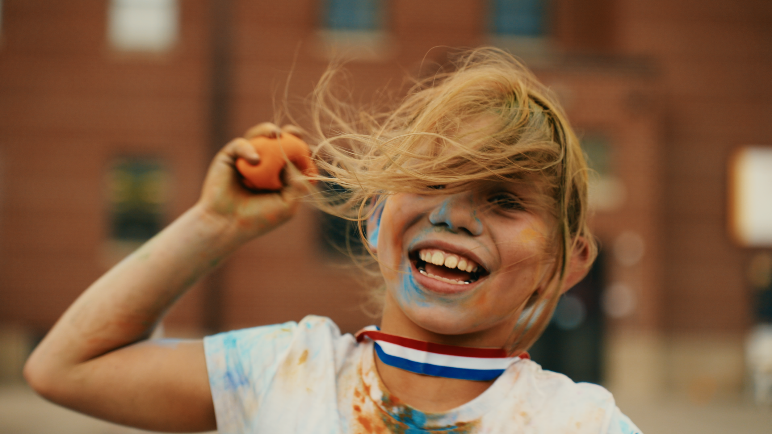 A young child with messy blond hair is smiling widely, holding a small orange object in their hand. Their face and arms are covered with colorful paint, and they appear to be enjoying a playful outdoor activity.