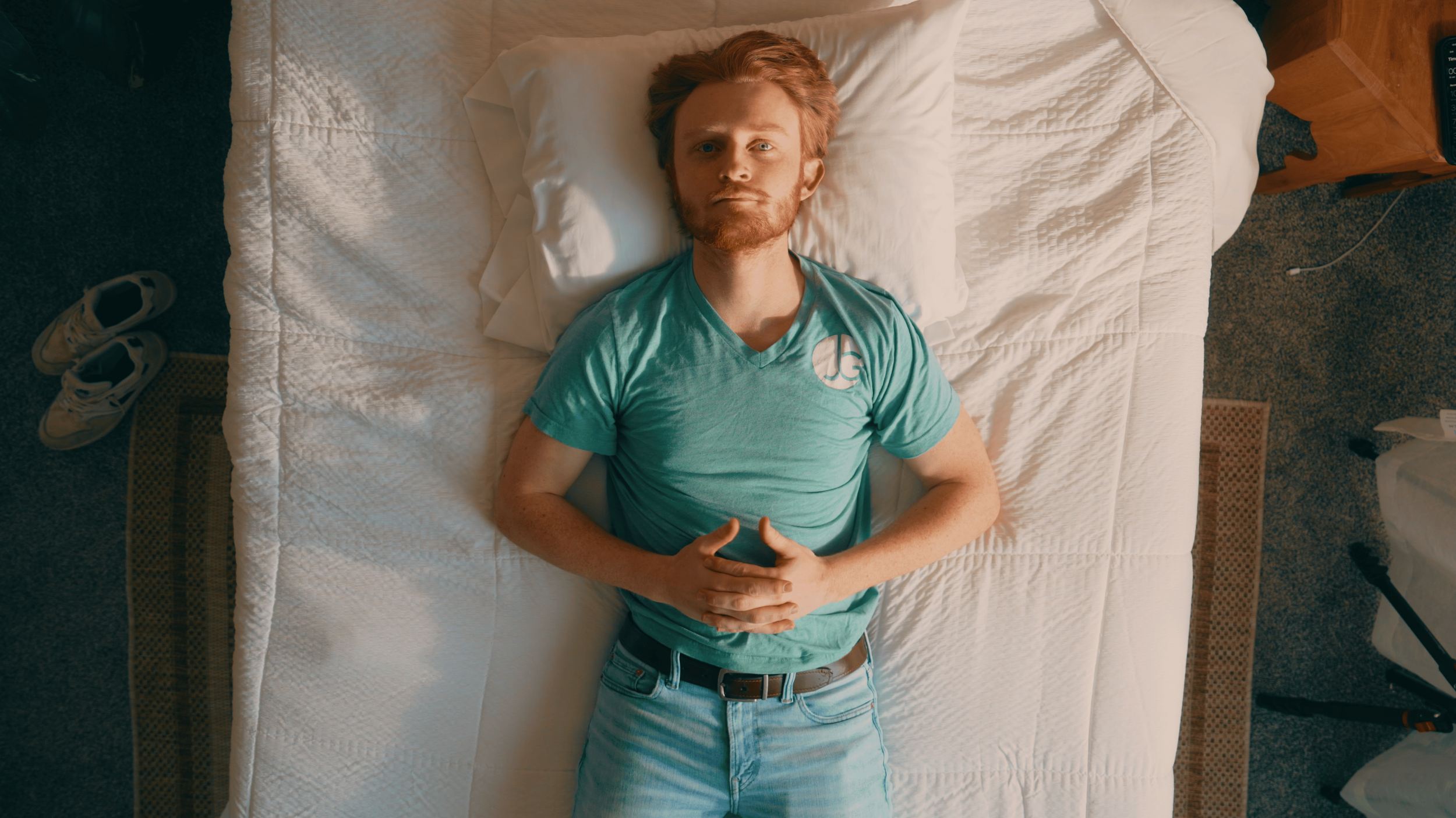 A young man with red hair and beard lying on a bed, looking up at the ceiling, wearing a turquoise t-shirt and jeans.