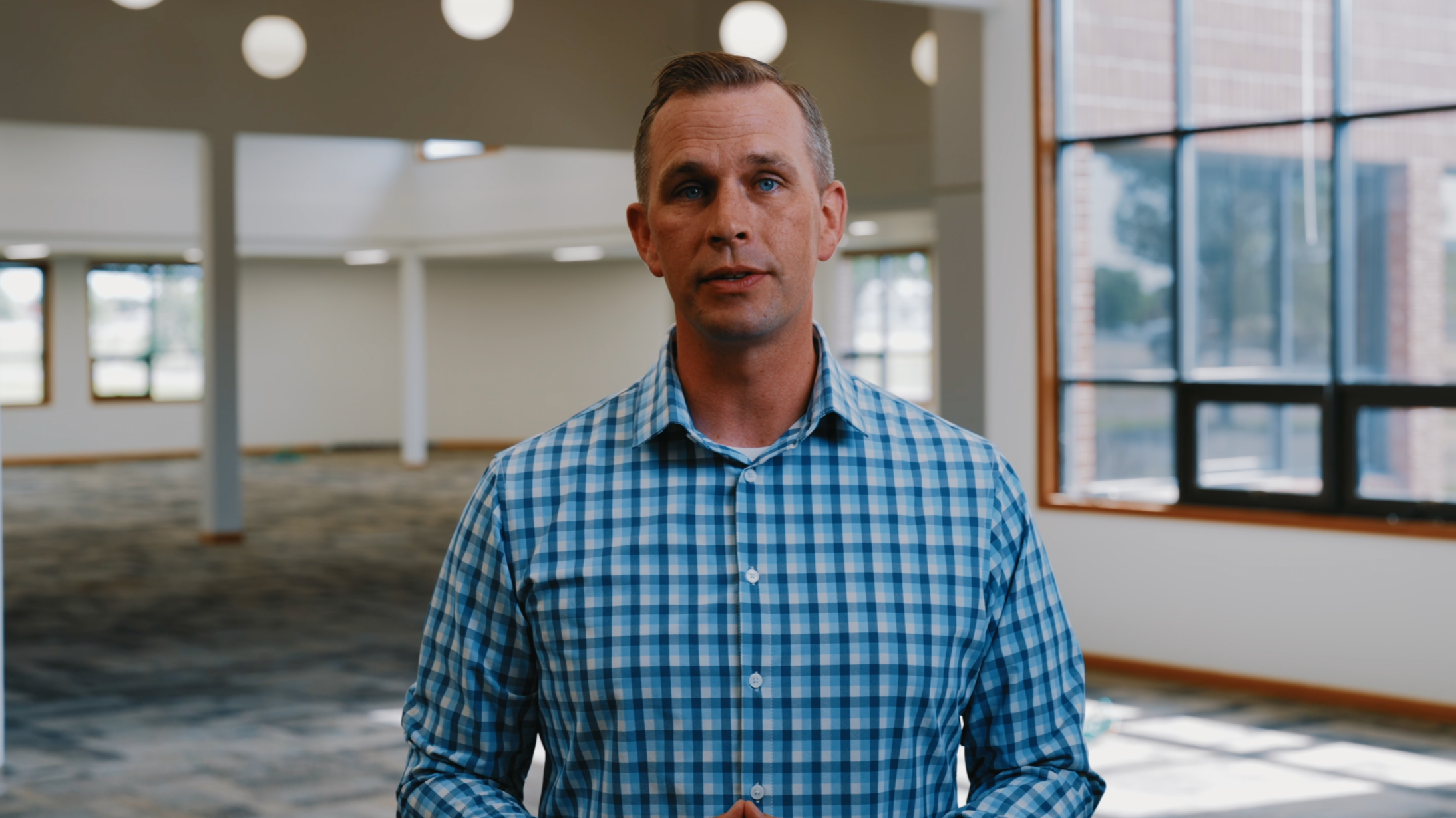 A man with short, light brown hair, blue eyes, and wearing a blue and white checkered shirt, standing indoors near large windows in a spacious, well-lit room.