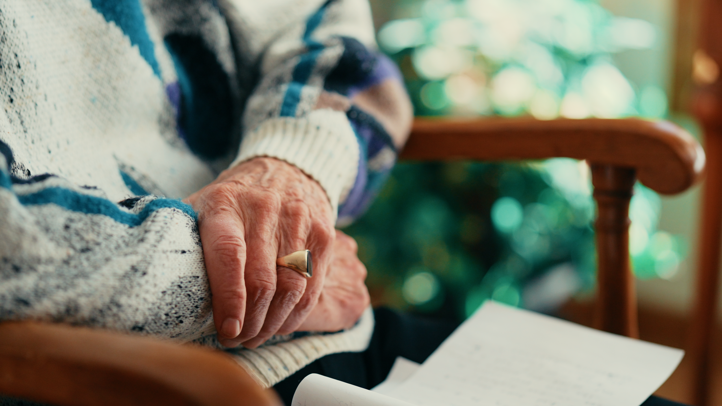 Close-up of an elderly person's hands resting on a table, wearing a patterned sweater. In the background, there is a wooden chair and blurred greenery with bokeh lights.