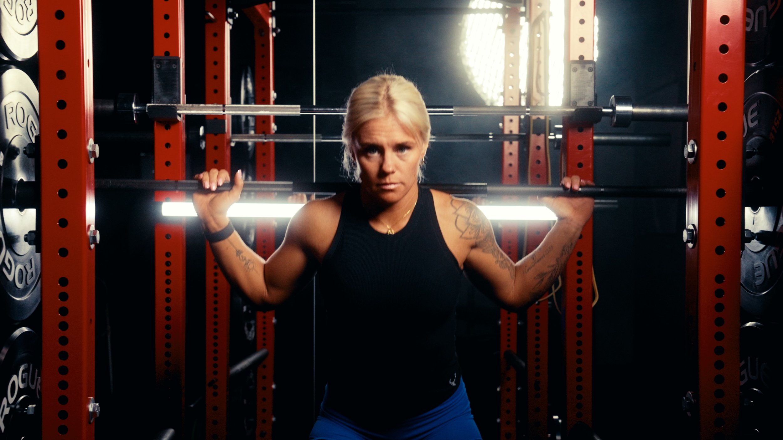 A woman with blonde hair and tattoos on her arms holding a barbell on her shoulders in a gym with red workout equipment and weight plates in the background.