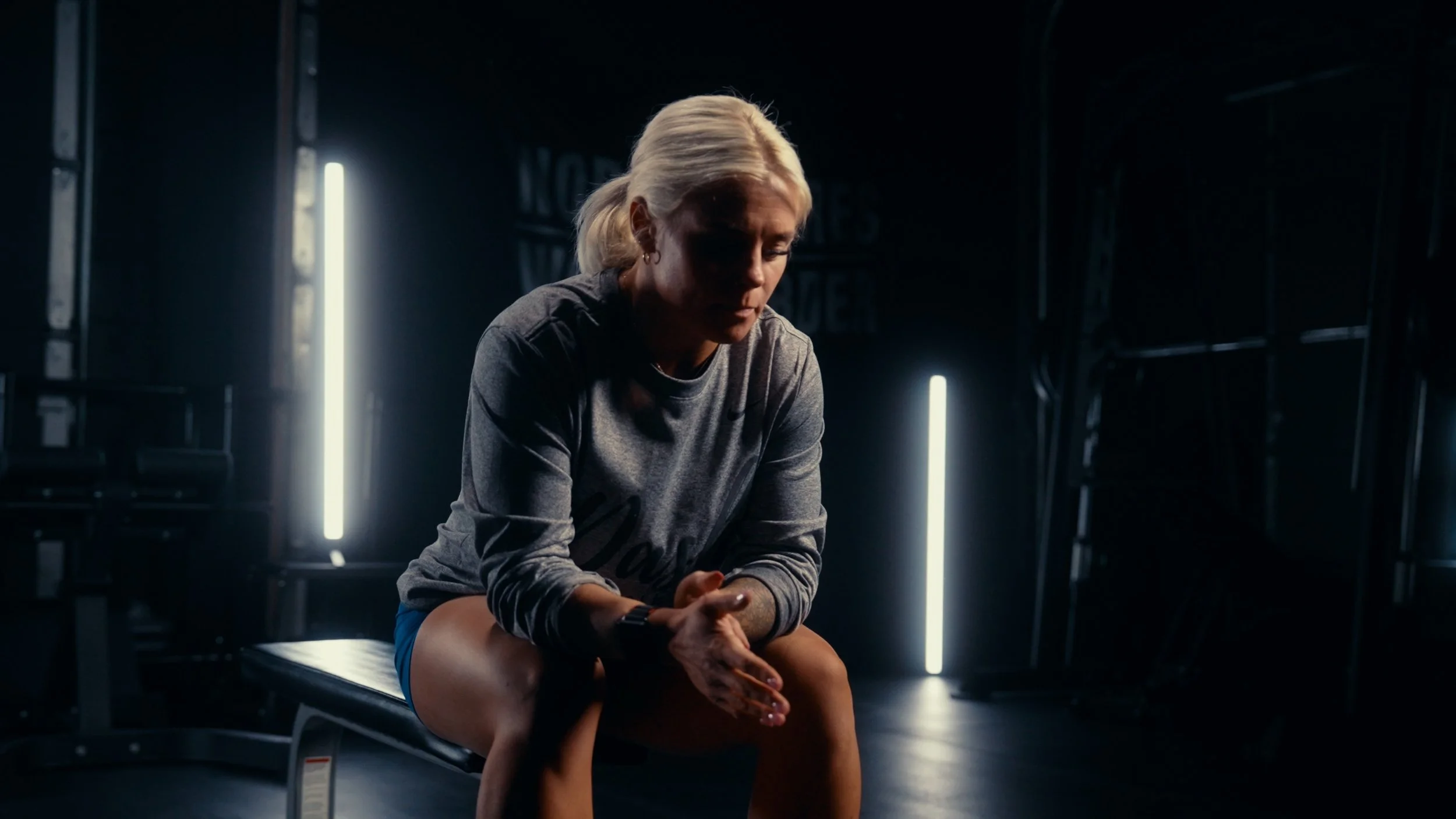 A woman with blonde hair sitting on an examination table in a dimly lit room with vertical white lights behind her, looking down with a contemplative expression.