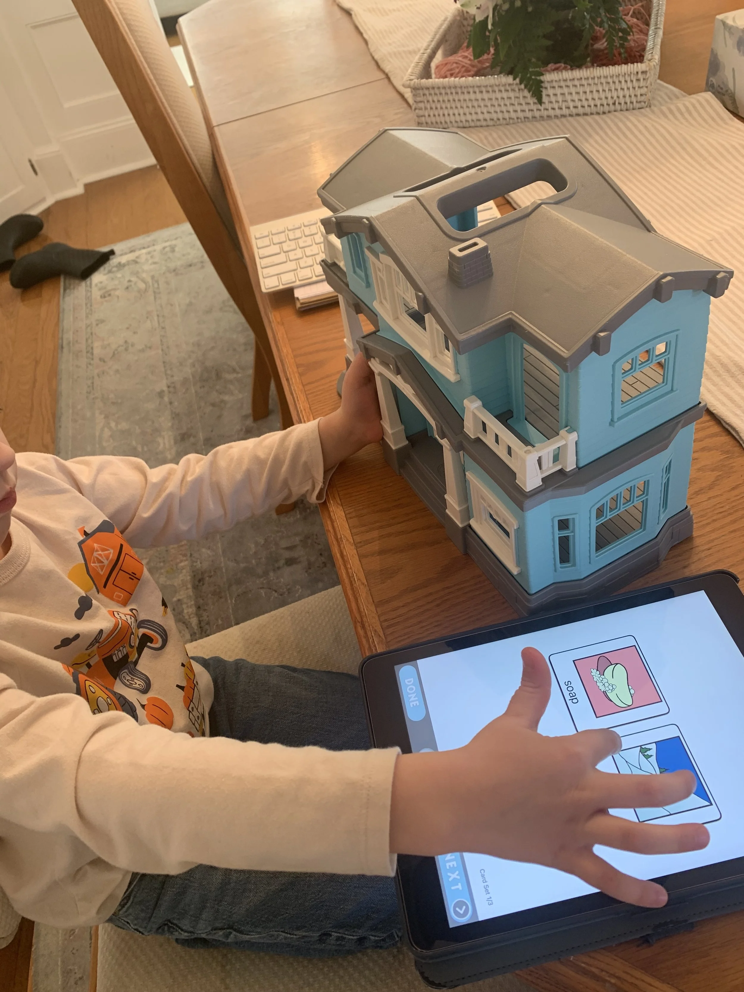 Child playing with a dollhouse on a dining table, with a tablet showing a picture of soap and other images.