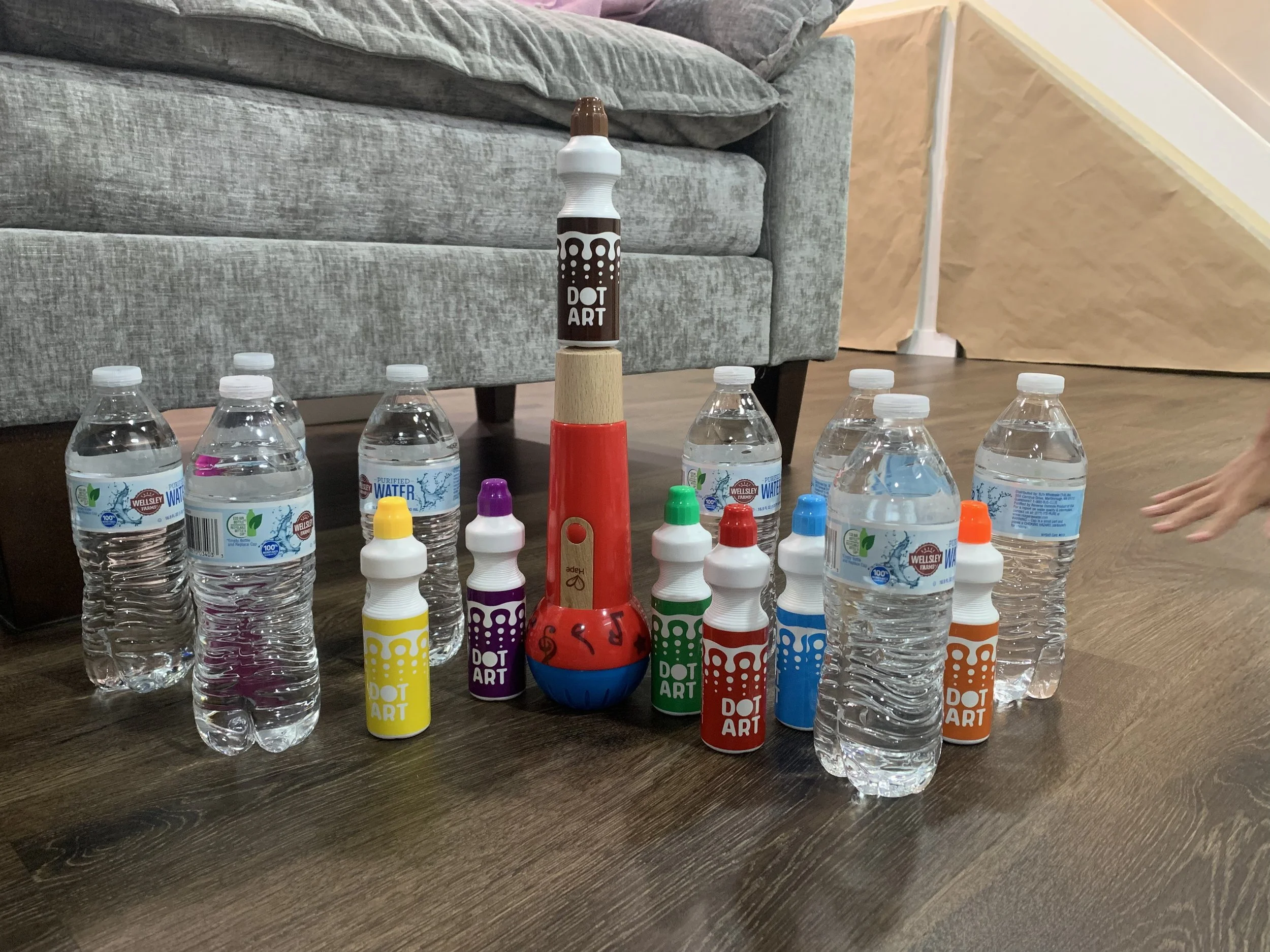 Multiple plastic water bottles and colorful dot art paint bottles arranged on a wooden floor, with a gray sofa and a beige wall in the background.