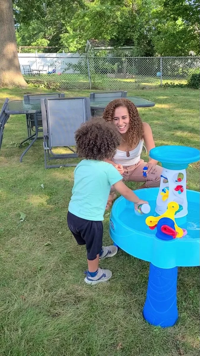 A woman and a young boy playing with a water toy in a backyard on a sunny day, with trees, a fence, and outdoor furniture in the background.