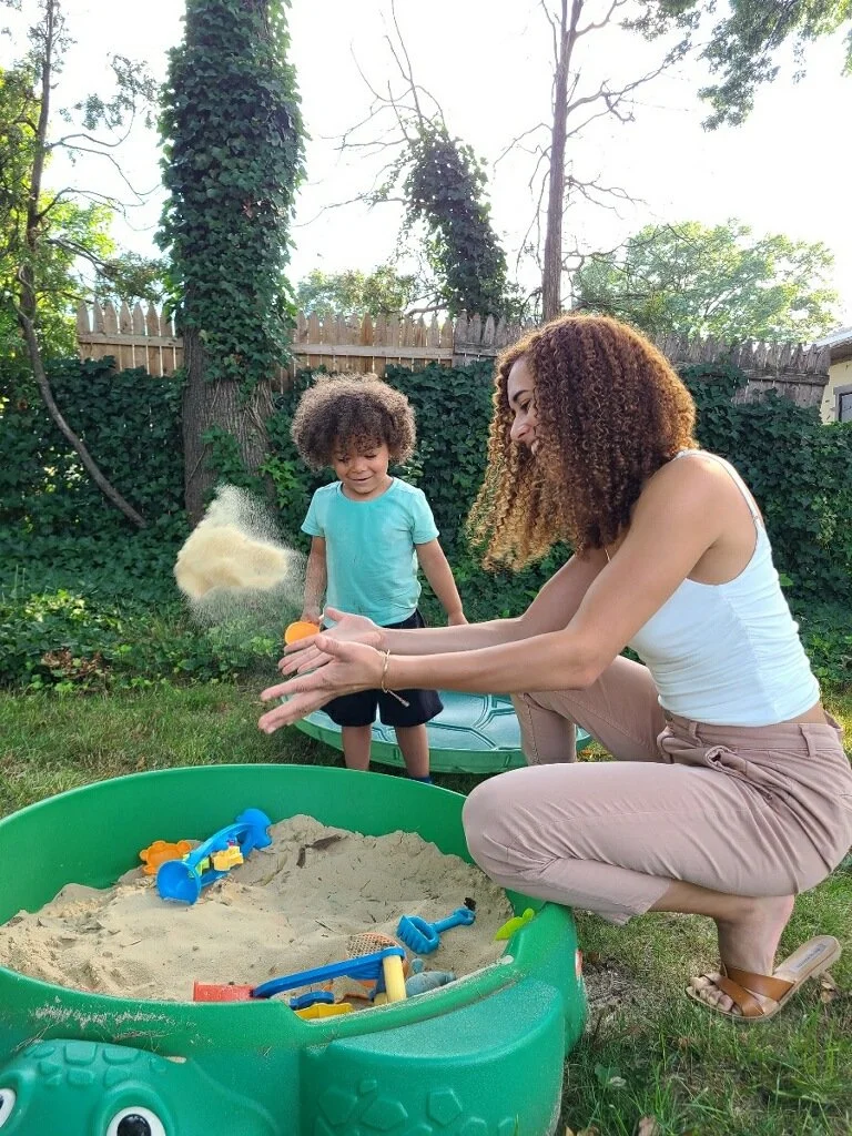 A woman and a young boy playing with a sandbox in a backyard. The woman is sitting, wearing a white tank top and beige pants, while the boy stands nearby. They are surrounded by greenery and trees, with a wooden fence in the background. The sandbox contains various colorful toy shovels and a sandcastle