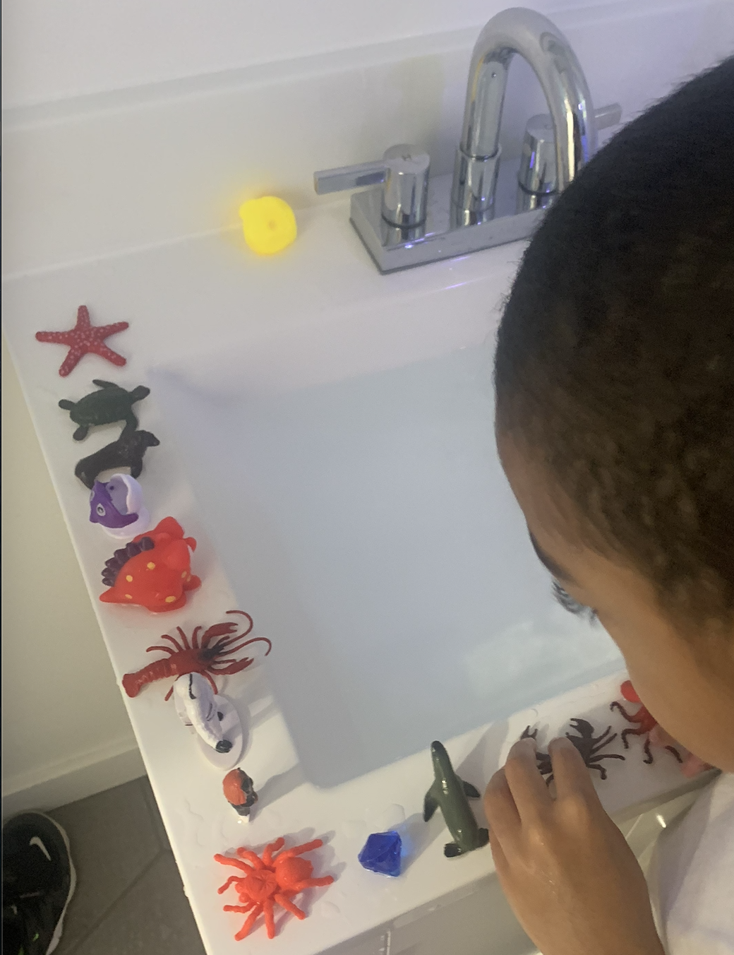 Child arranging toy sea creatures on a white surface near a sink with chrome faucet, featuring a starfish, turtle, purple fish, crab, and other sea animal toys.