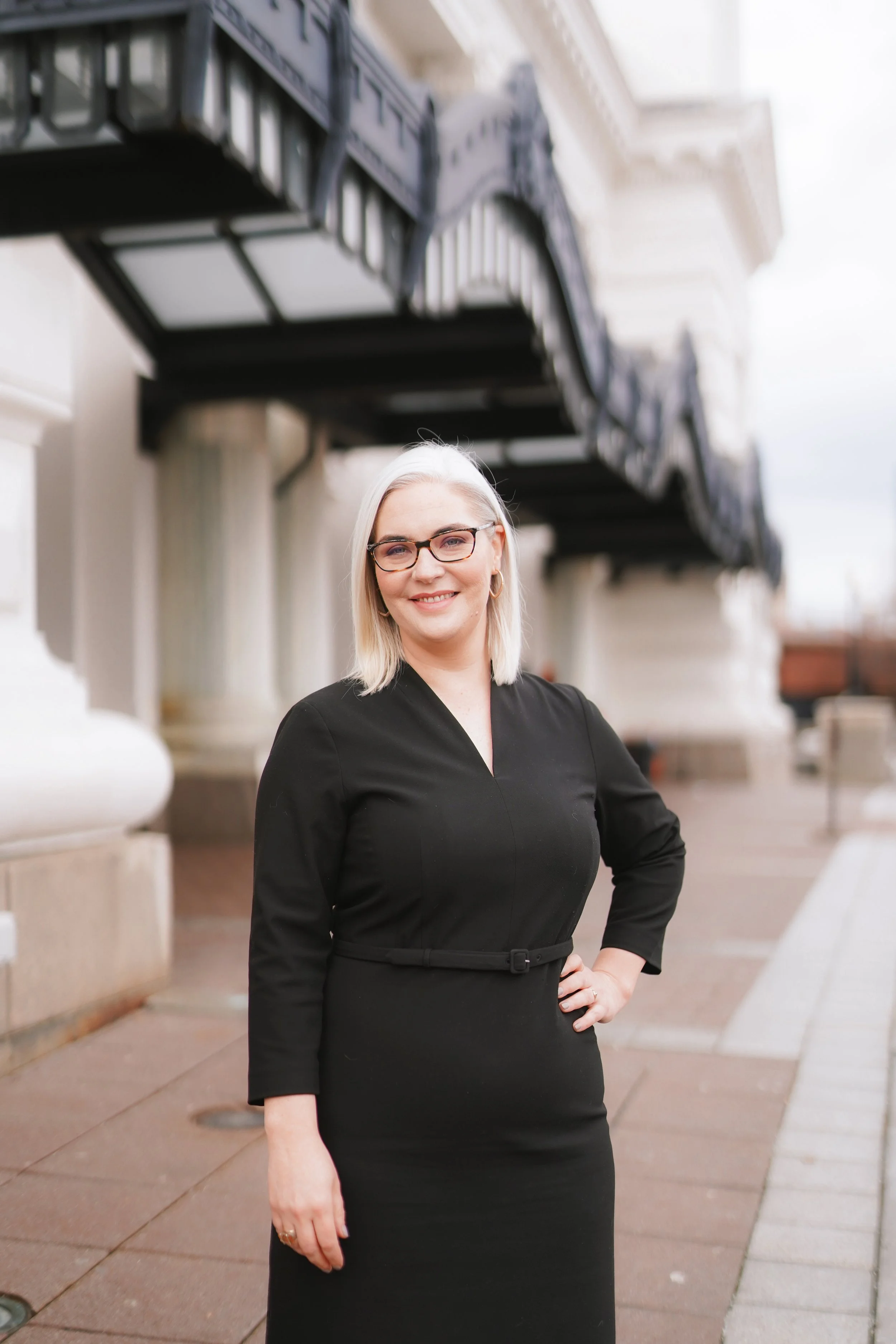 A woman with blonde hair, glasses, and earrings, wearing a black dress, standing outdoors in front of a building with ornate architecture and black balconies, smiling at the camera.