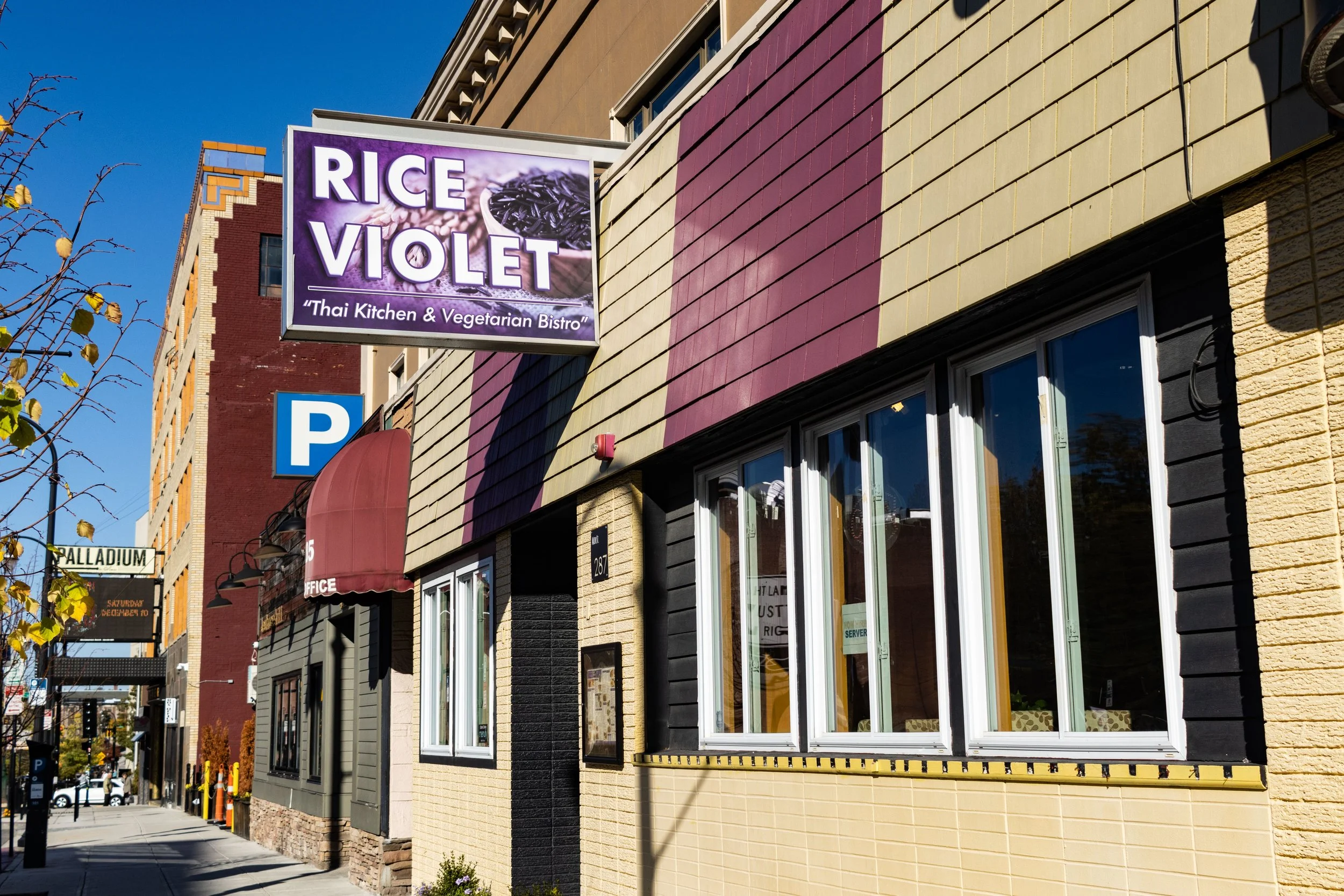 Street view of Rice Violet Thai Kitchen & Vegetarian Bistro with a purple and yellow striped exterior, a maroon awning, and a sign displaying the restaurant's name. There's a parking sign and nearby retail building in the background.