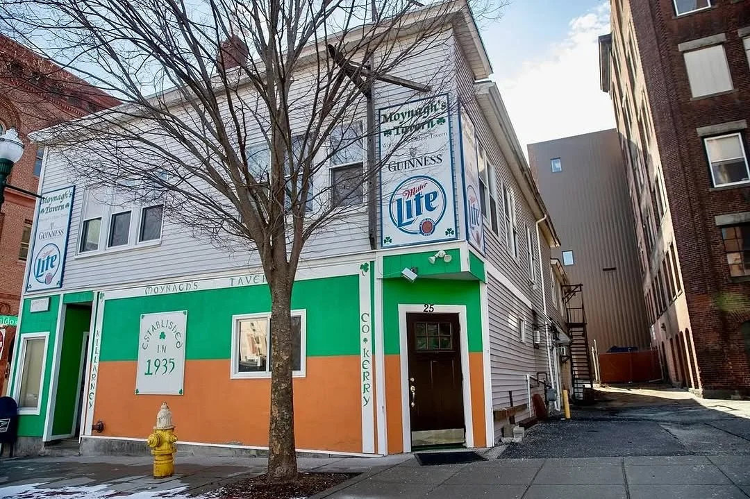 Street view of a two-story building with a white and green exterior, featuring a sign that indicates it is a bakery established in 1935, with advertisements for Miller Lite and Guinness beer. There is a tree in front, and an alleyway to the right.