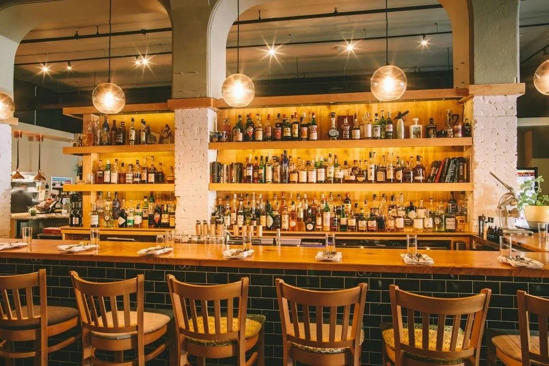 Bar with a variety of liquor bottles on wooden shelves behind the counter, illuminated by warm lighting and modern spherical pendant lights, with wooden chairs at the bar.