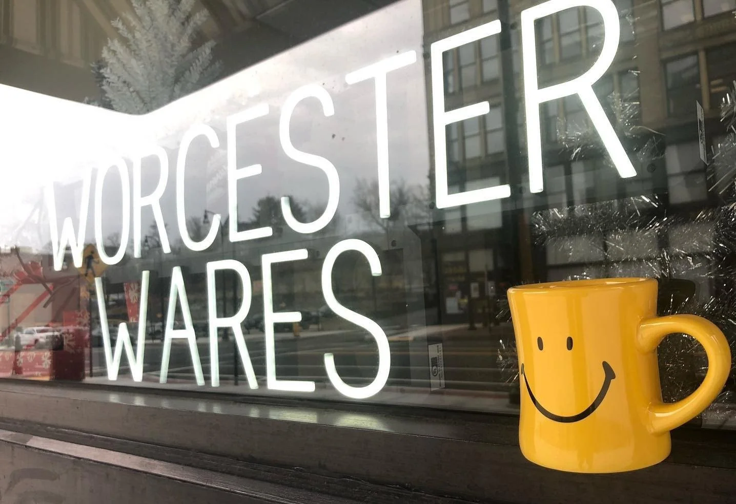 A storefront window with a neon sign reading 'Worcester Wares' and a yellow smiling mug placed on the window sill, with reflections of trees and buildings visible in the window.