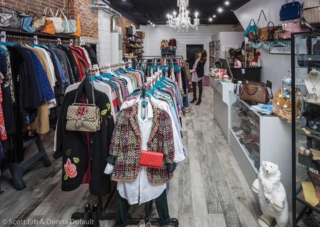Interior of a clothing boutique with racks of various garments, handbags on display, and a glass display case filled with accessories. There is a chandelier hanging from the ceiling and a white bear statue next to the counter.