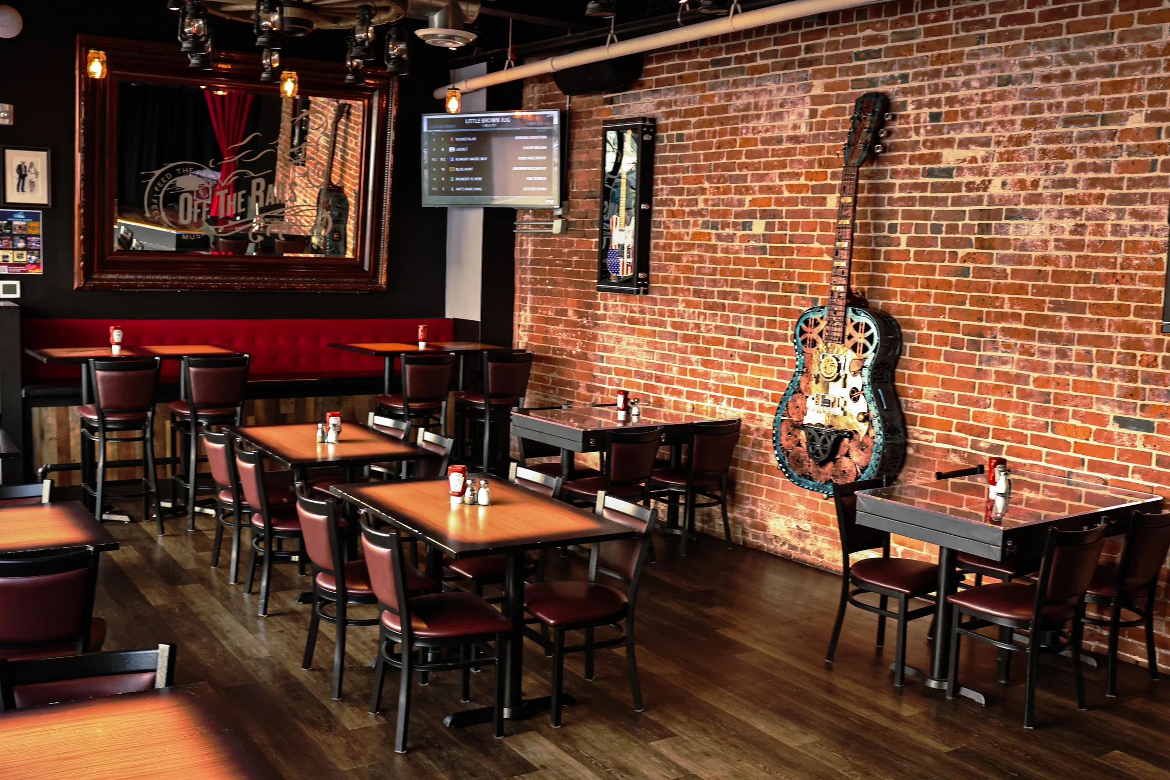Interior of a music-themed restaurant or bar with brick walls, several wooden tables with dark chairs, a high red cushioned bench, a large mirror with the text 'Off the Record,' a mounted TV screen, and guitar decor on the wall.