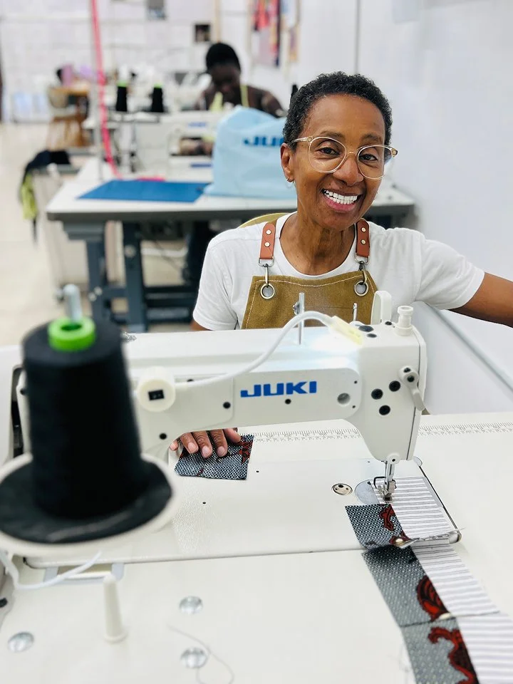 Smiling woman with glasses working on a sewing machine in a textile workshop.