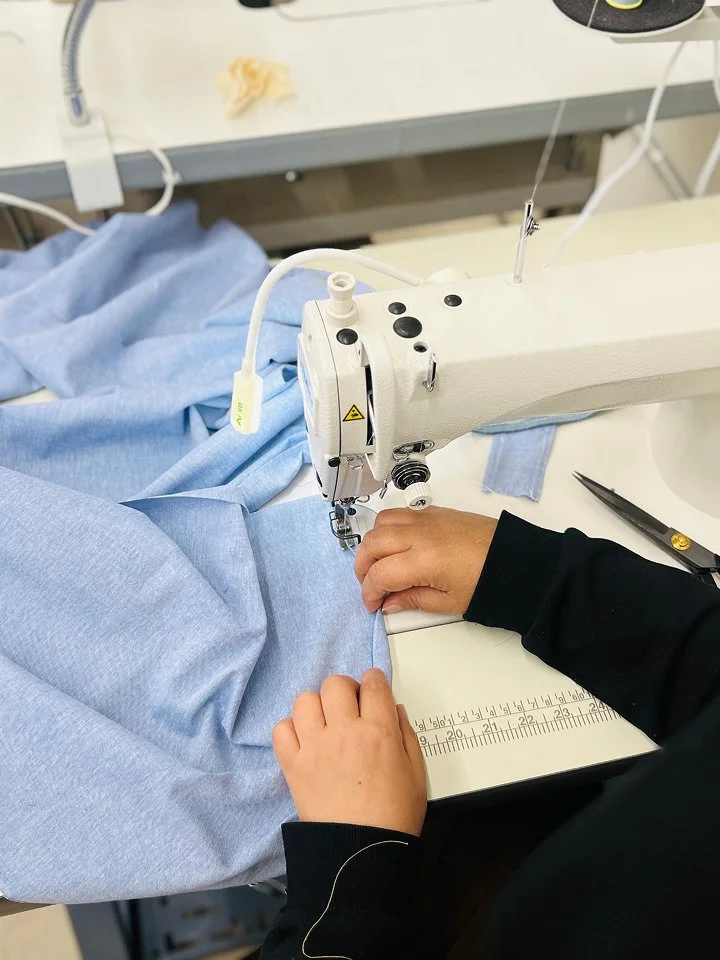 Person sewing light blue fabric on a sewing machine, with scissors and yellow fabric piece nearby, on a white table in a clinical or workshop setting.