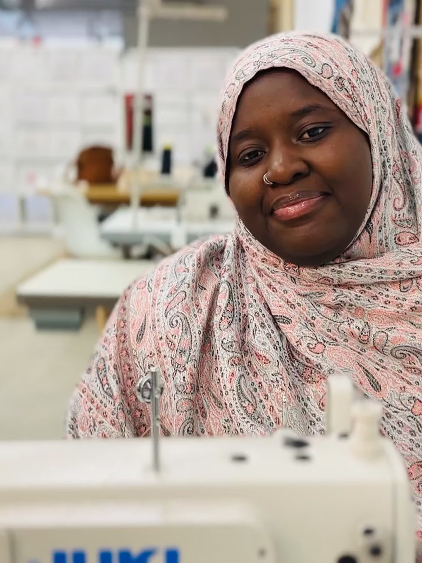 A woman wearing a pink patterned hijab sits in front of a sewing machine in a room that appears to be a sewing or craft workshop, with shelves and supplies in the background.