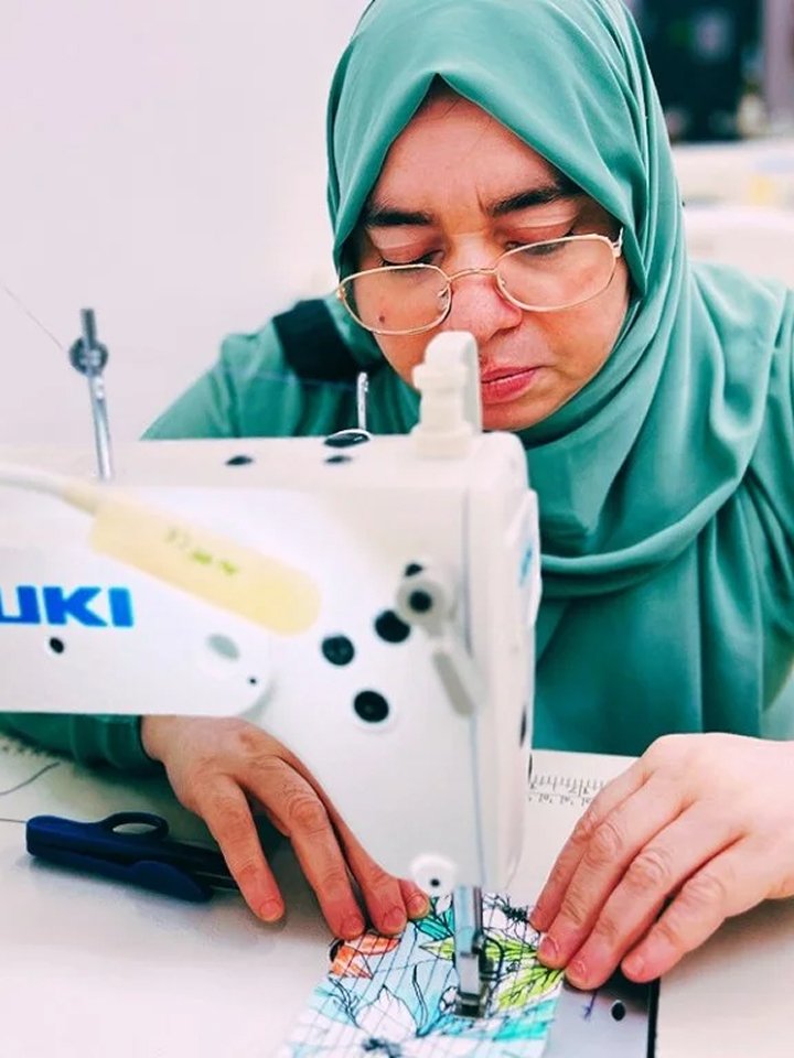 A woman wearing a green hijab and glasses sewing fabric with a white sewing machine.