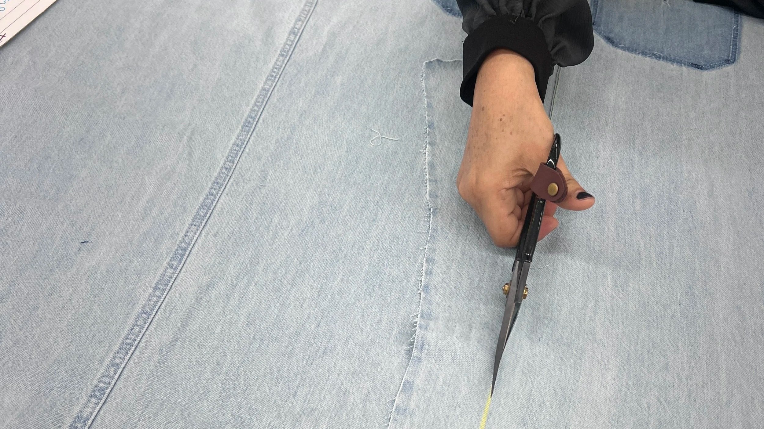 A person in black gloves is using scissors to cut a seam in light blue denim fabric.