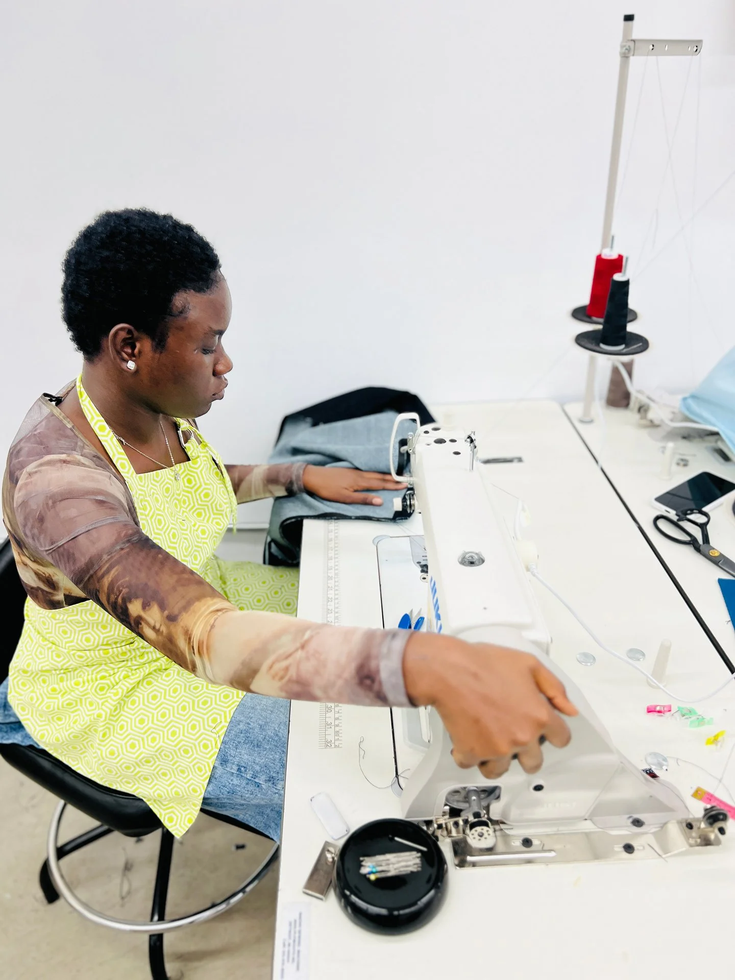 A woman working on a sewing machine in a well-lit room, wearing a yellow apron and earrings, with supplies and thread spools nearby.