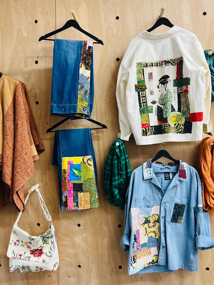 Clothing display on a wooden wall featuring embroidered denim jeans, a white jacket with a colorful Asian-inspired design, a light blue button-up shirt with patches, and a floral bag.