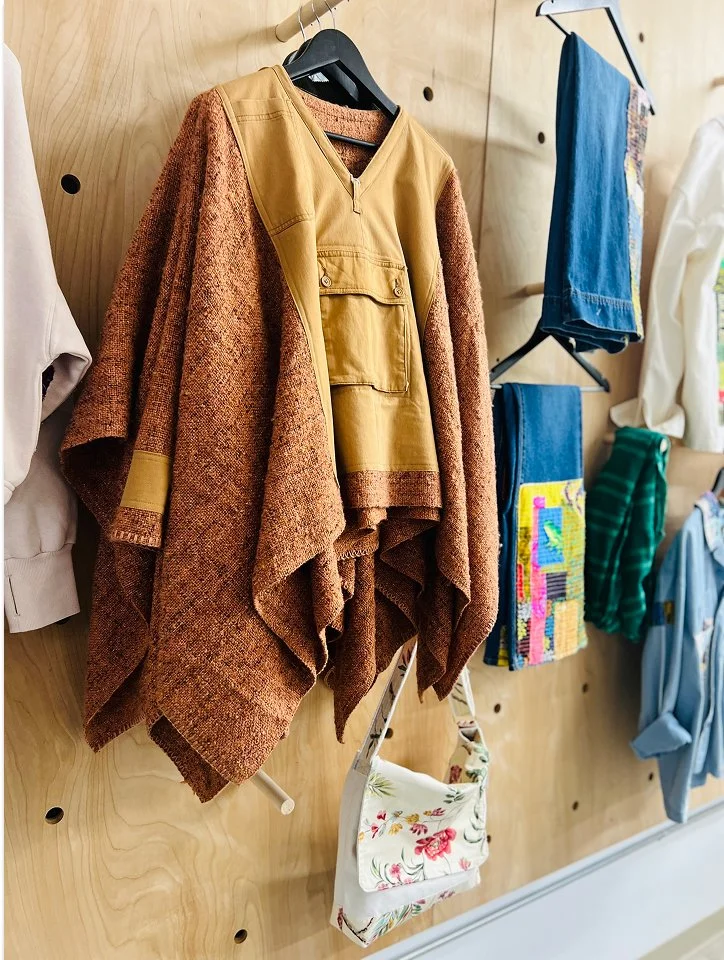 A displayed brown and mustard-colored cape with a matching vest and a white floral purse hanging from it, surrounded by other clothing and fabric items on a wooden wall.