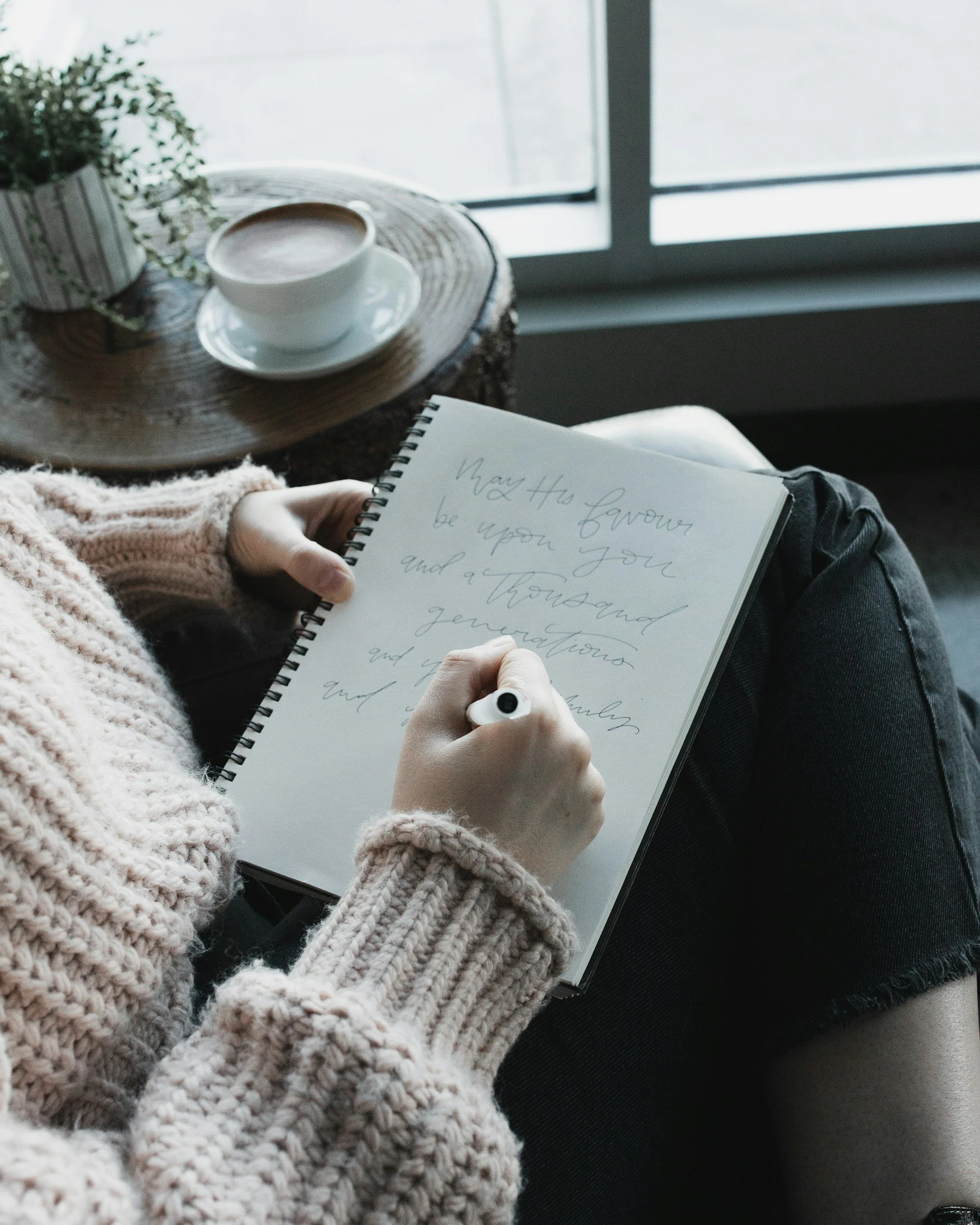 A person in a thick beige sweater holding a spiral notebook with handwritten text, a white ceramic cup and saucer with coffee on a rustic wooden table, next to a potted plant, sitting by a window with natural light.