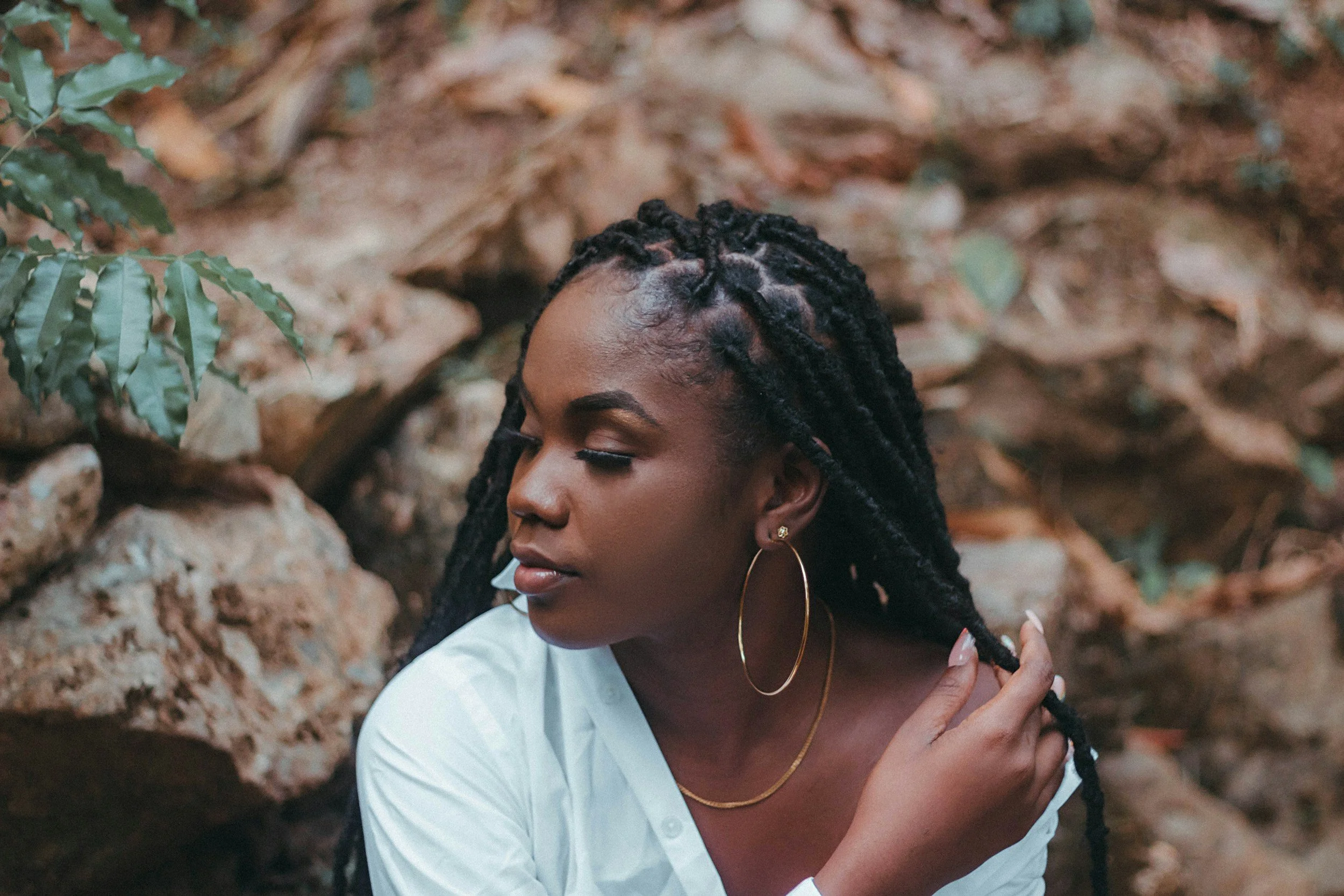 A woman with dark skin and long dreadlocks, wearing hoop earrings and a white shirt, is seen in profile against a background of rocks and dry leaves. She has her eyes closed and is touching her hair.