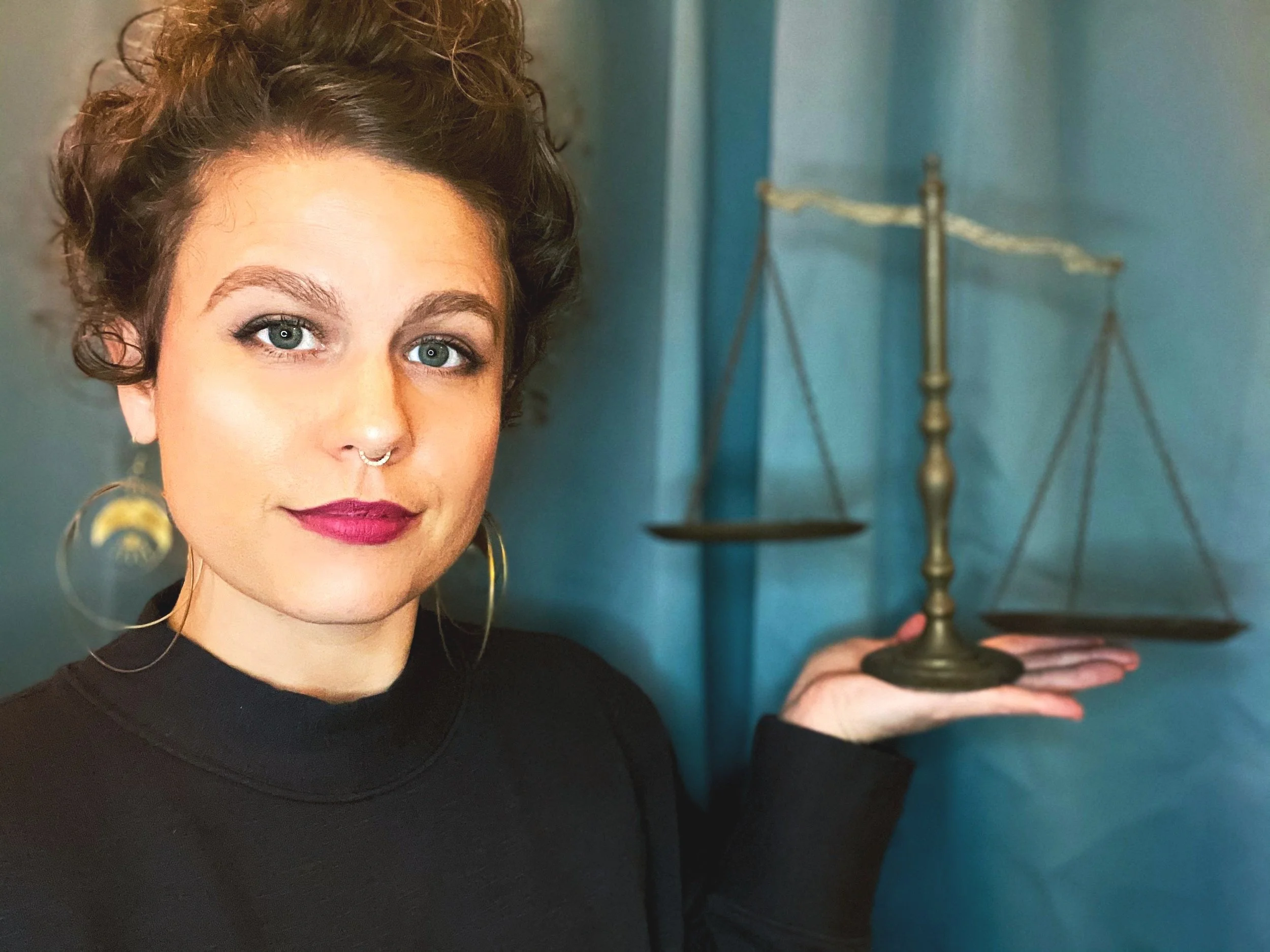 A person with short curly hair, wearing a black top, large hoop earrings, and a septum piercing, is holding a brass balance scale in her right hand. She has bright eyes and bold lipstick, standing against a teal wall.