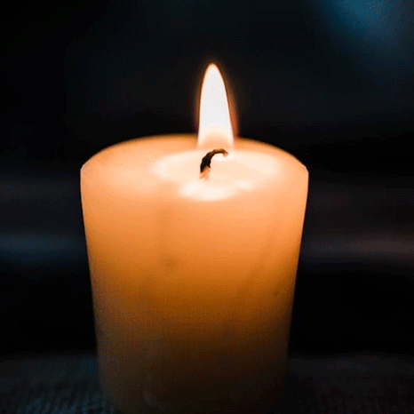 A lit beige-colored candle burning with a small flame against a dark background.