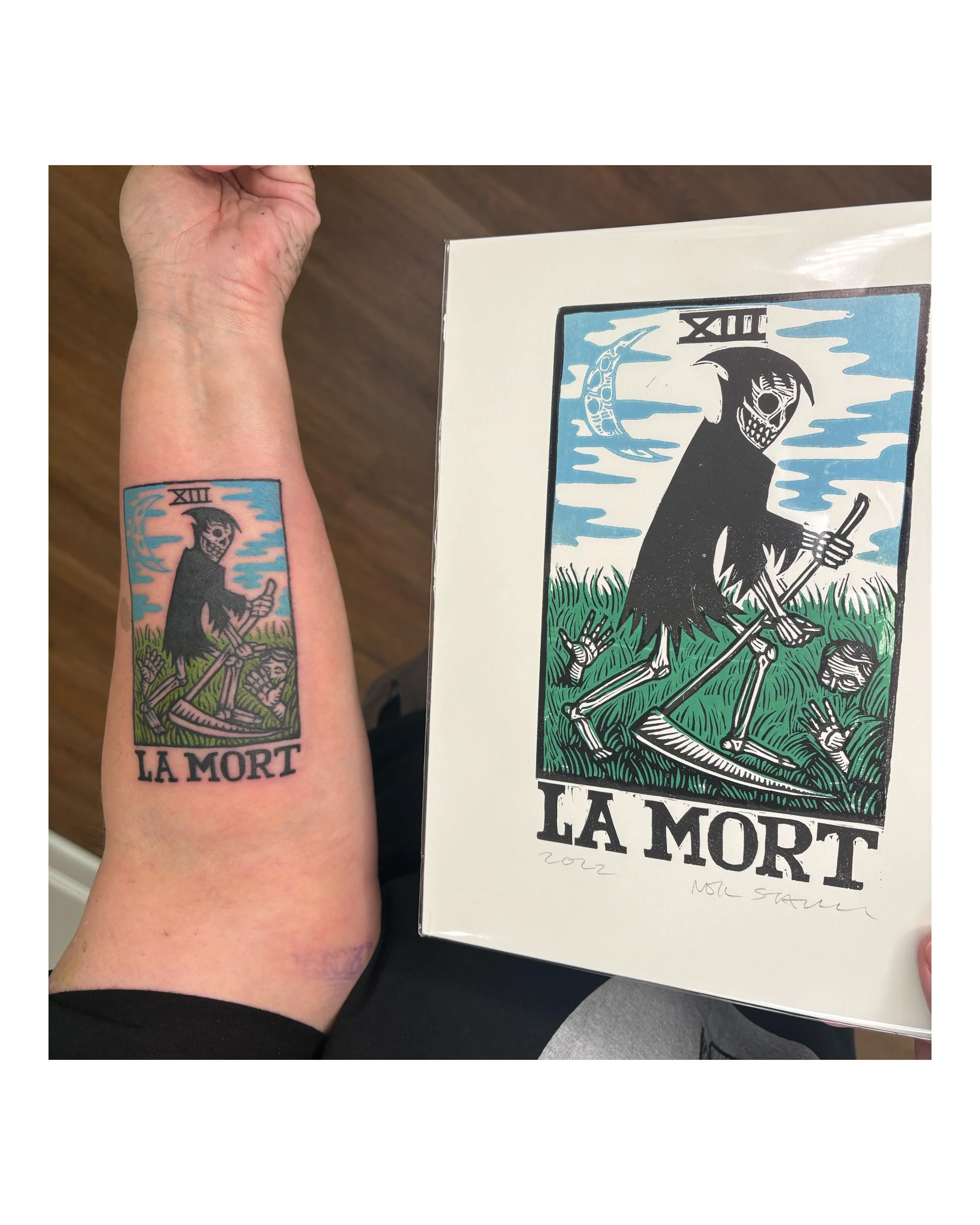 A tattoo on a person's forearm depicts a cartoonish skeletal figure dressed as the Grim Reaper, holding a sickle, with the text "XIII LA MORT" underneath. The person is holding a box featuring the same artwork. Custom Art By Neil Stavely 