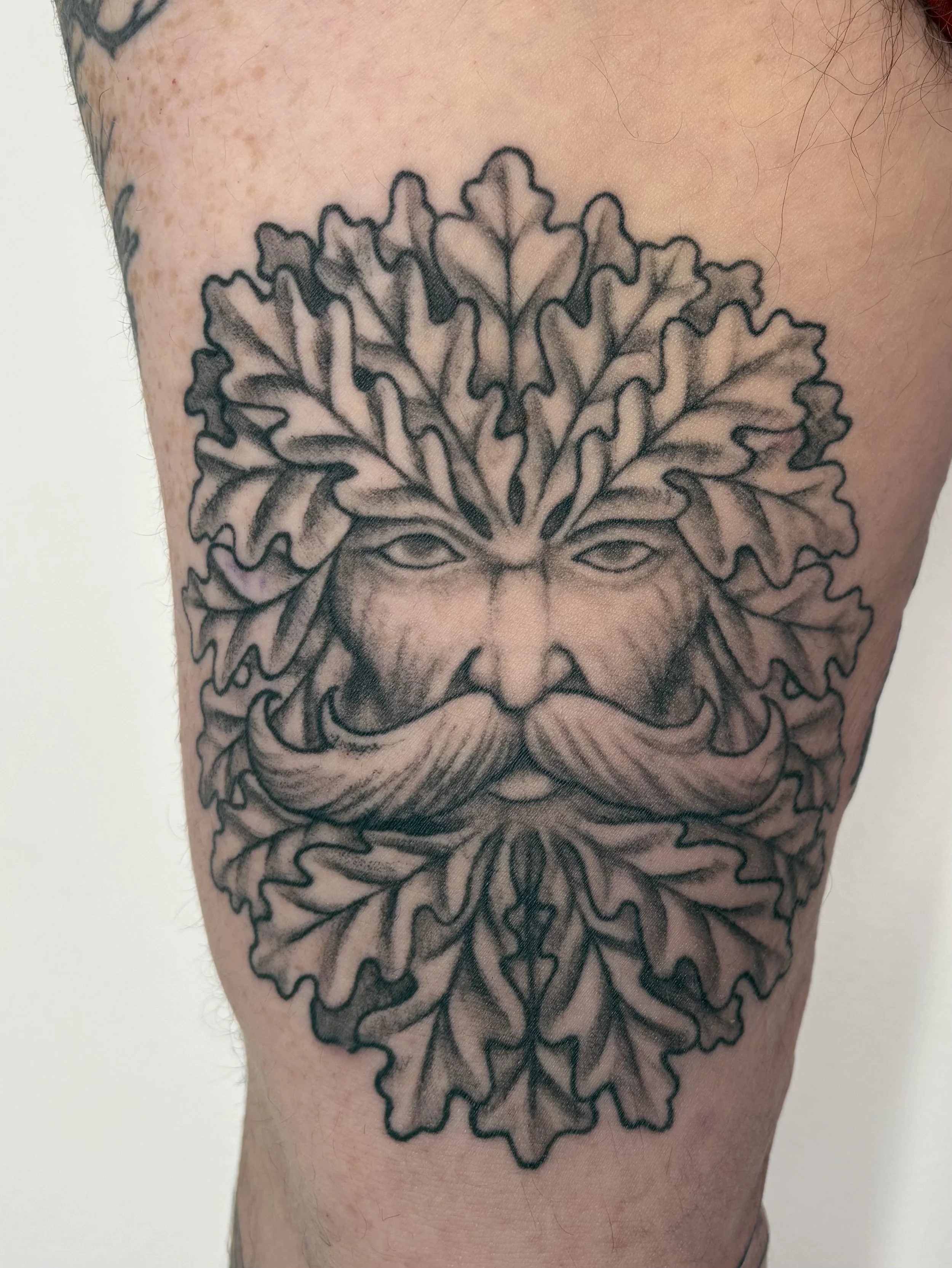 Tattoo of a face with oak leaves surrounding it on a person's upper arm. State Of The Art Tattooing Winchester Virginia 