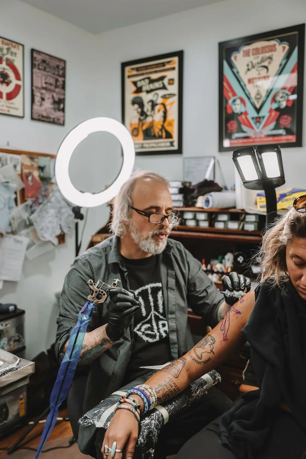 A tattoo artist is tattooing a woman's arm in a tattoo parlor with framed artwork on the walls and equipment on shelves.