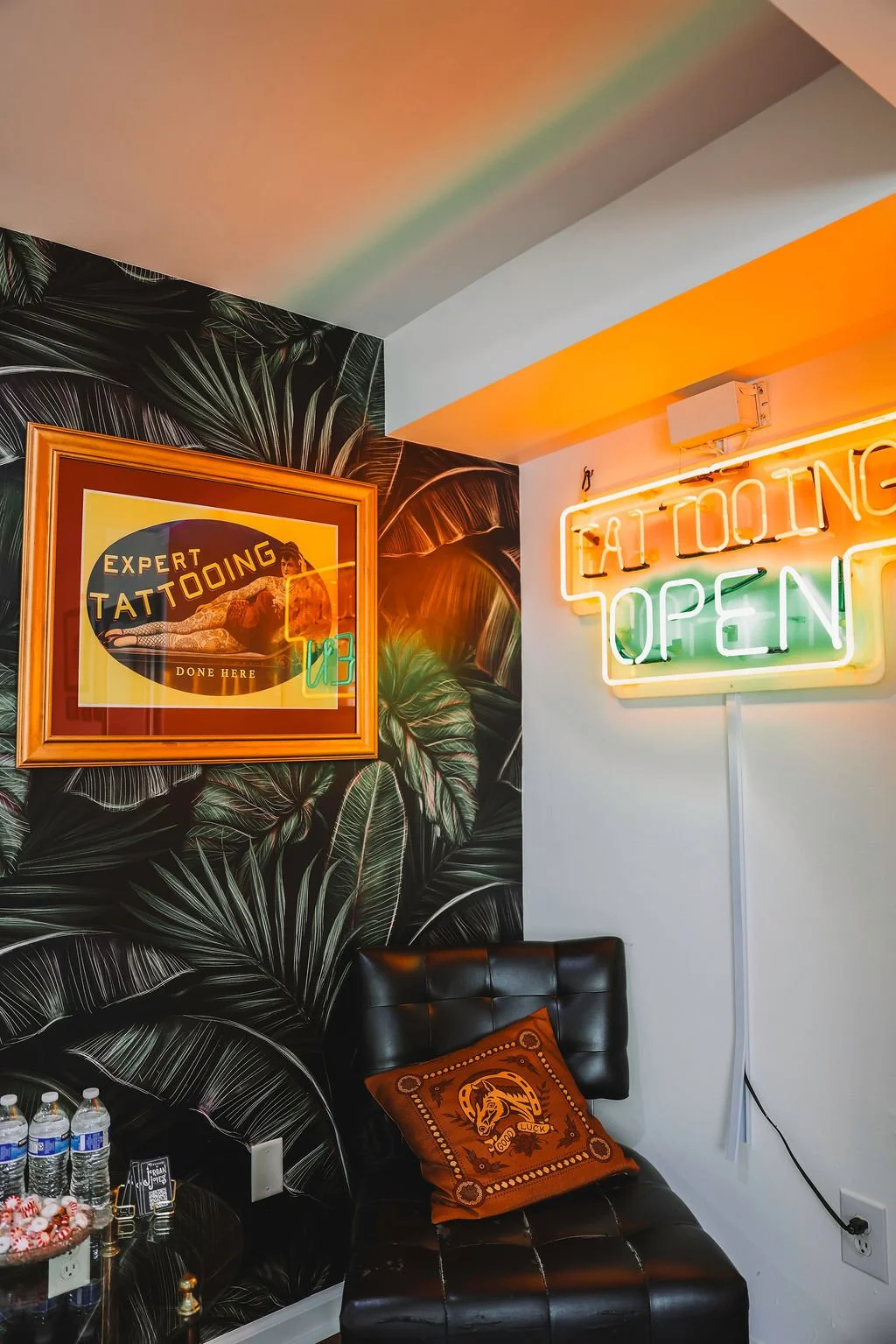 Interior of a tattoo parlor in Winchester Virginia , bottled water on a table, a framed poster advertising tattooing, and a neon sign that reads 'TATTOOING OPEN' with neon lights in orange, green, and yellow.