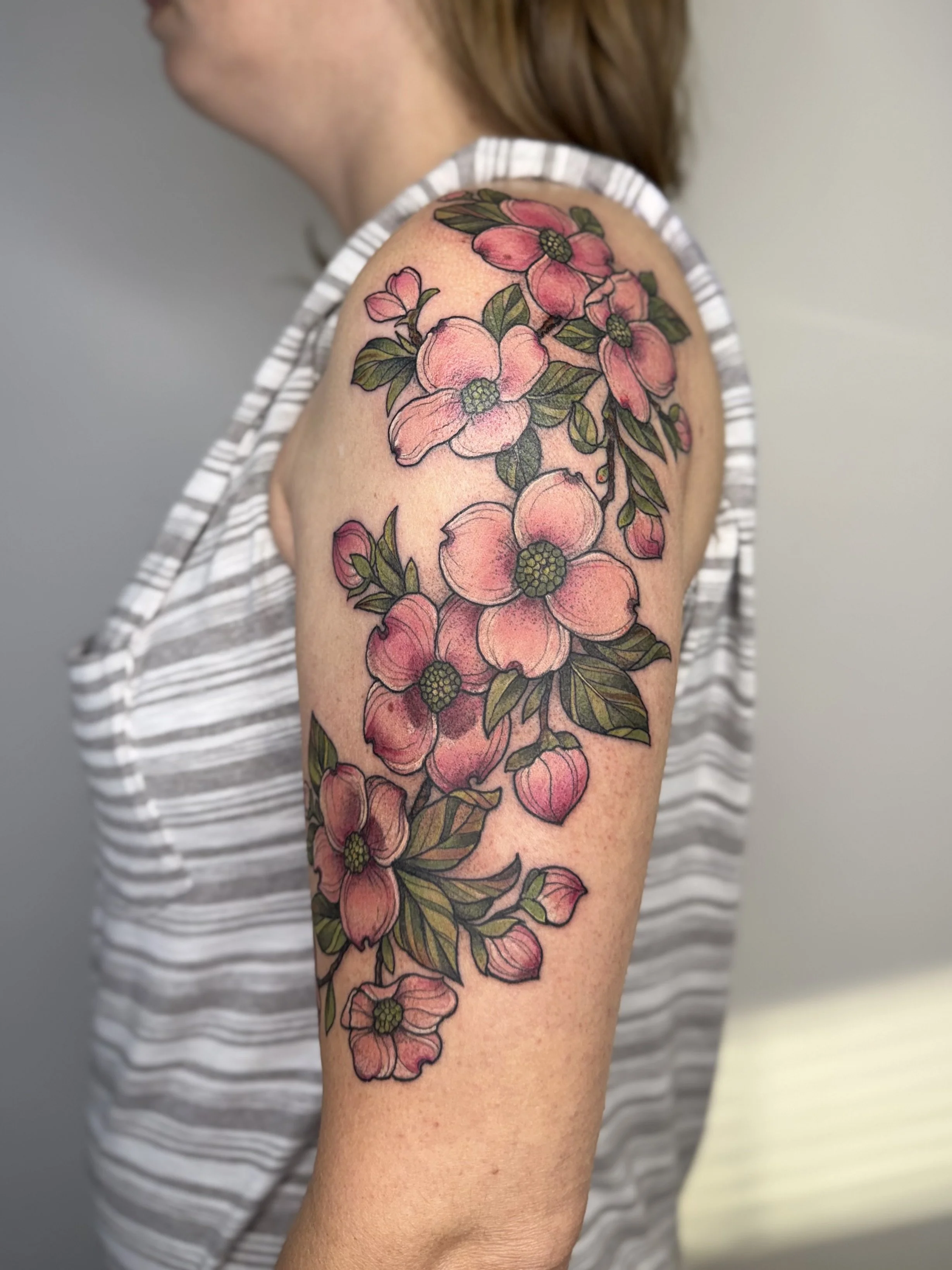 A person's upper arm and shoulder with a colorful floral tattoo featuring pink flowers with green leaves. The person is wearing a sleeveless striped top. Custom Tattoo by Jordan Joyes