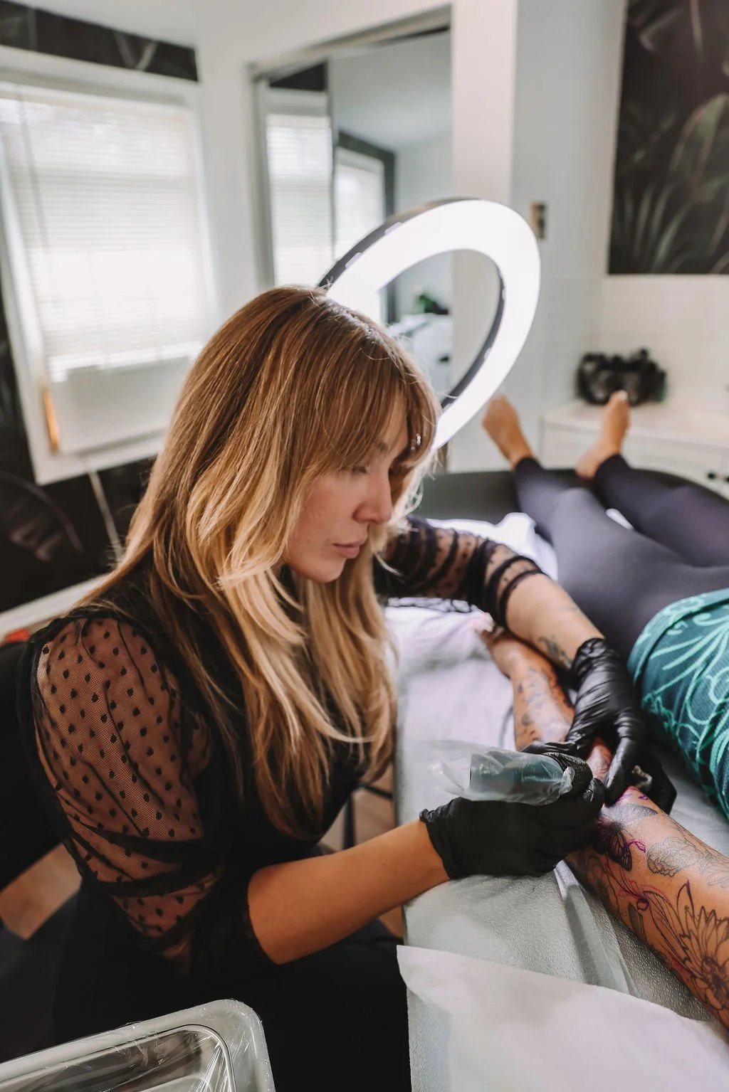 A woman in black gloves is tattooing a person's arm, which is covered with colorful tattoos, indoors with natural light and a ring light.