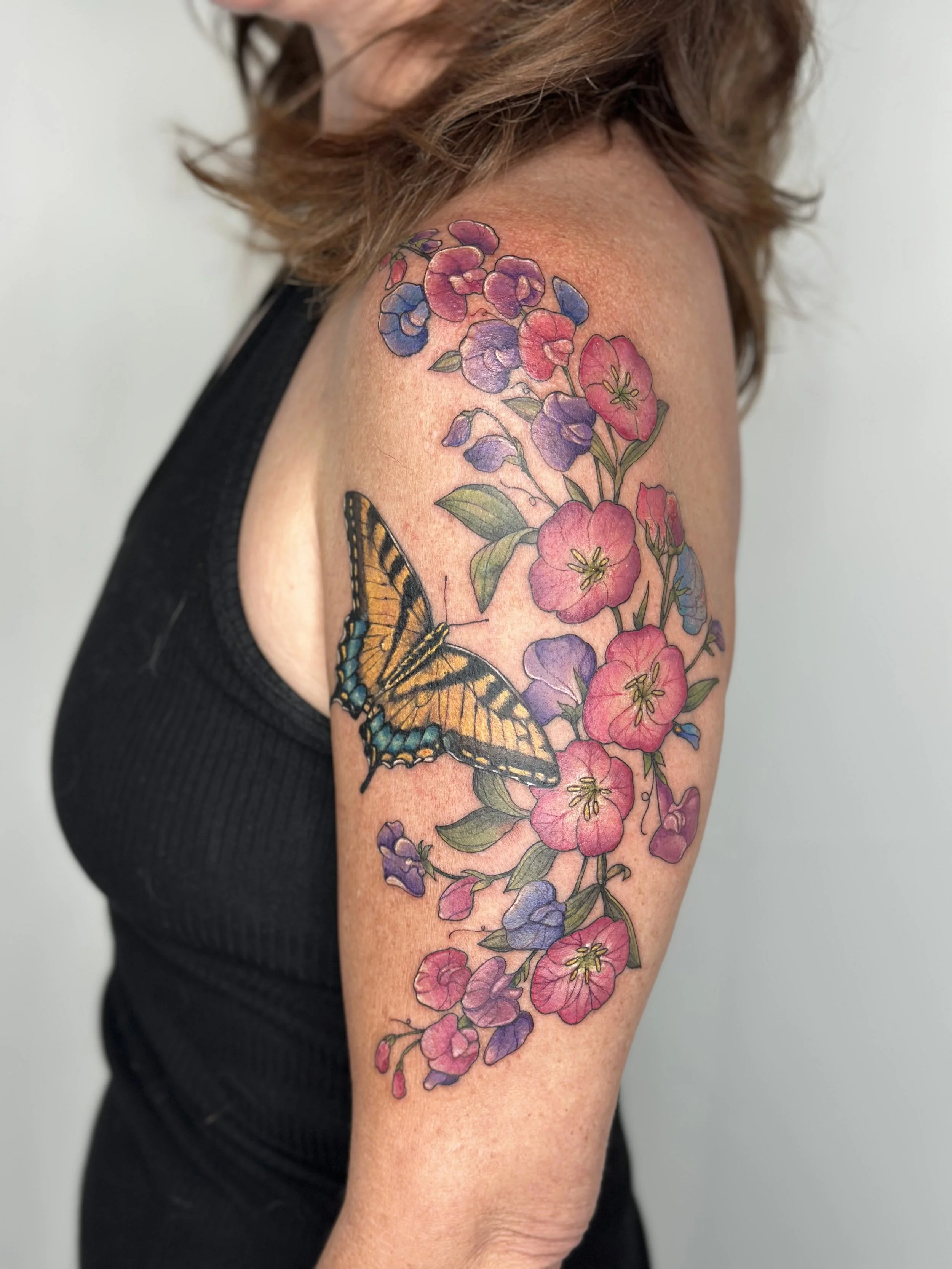 A person's shoulder and upper arm with a colorful tattoo by Jordan Joyes featuring pink and purple flowers, green leaves, and a large butterfly with orange, black, and blue markings. 