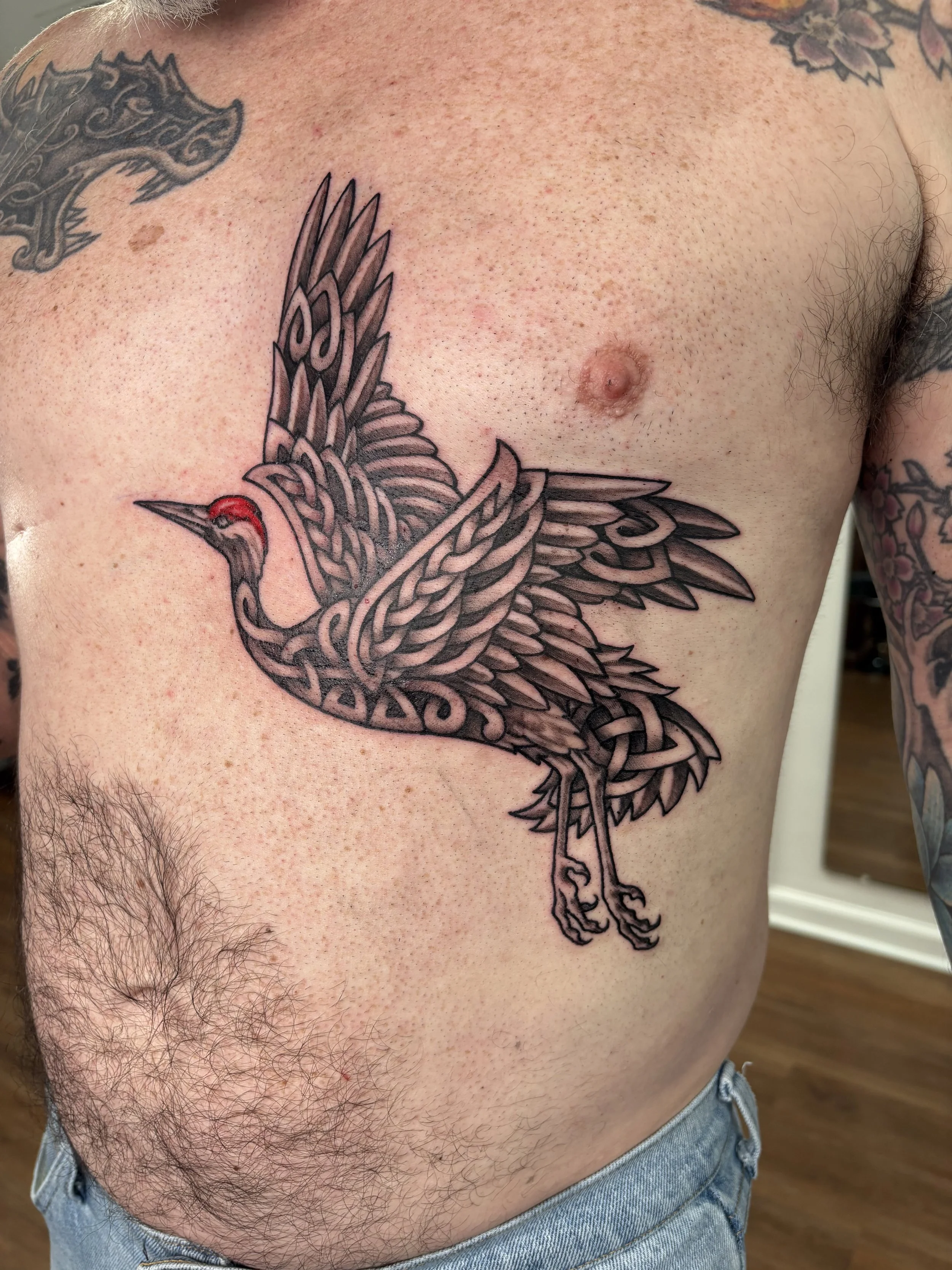 Tattoo of a stylized bird with intricate line work and red accents on the head, located on a man's side torso near the chest, with additional tattoos visible on the shoulder and arm.State of The Art Tattooing Winchester VA