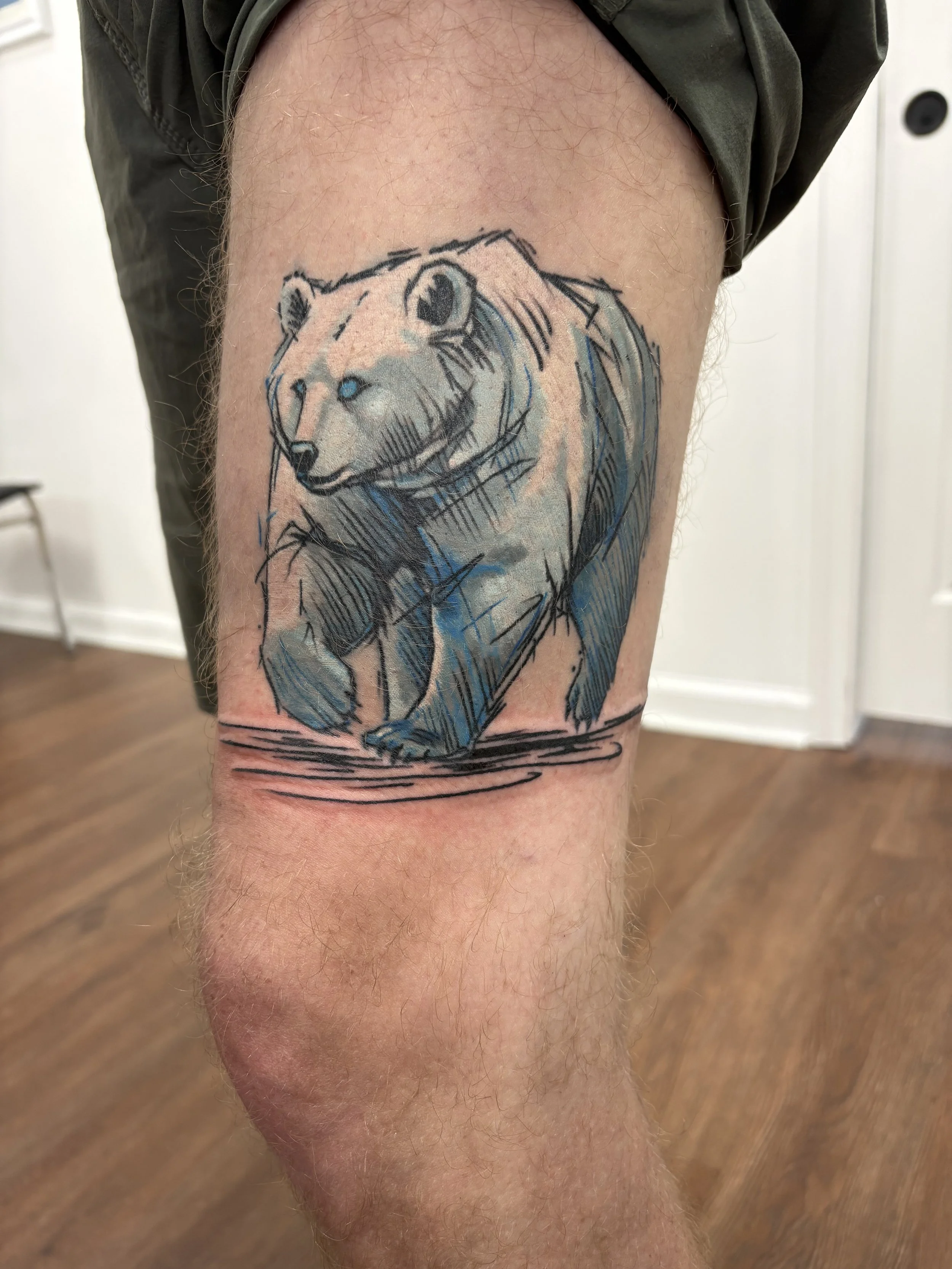 A tattoo of a lion's face on a person's thigh, with the lion's mane extending downward, rendered in sketch style with black ink and hints of blue.