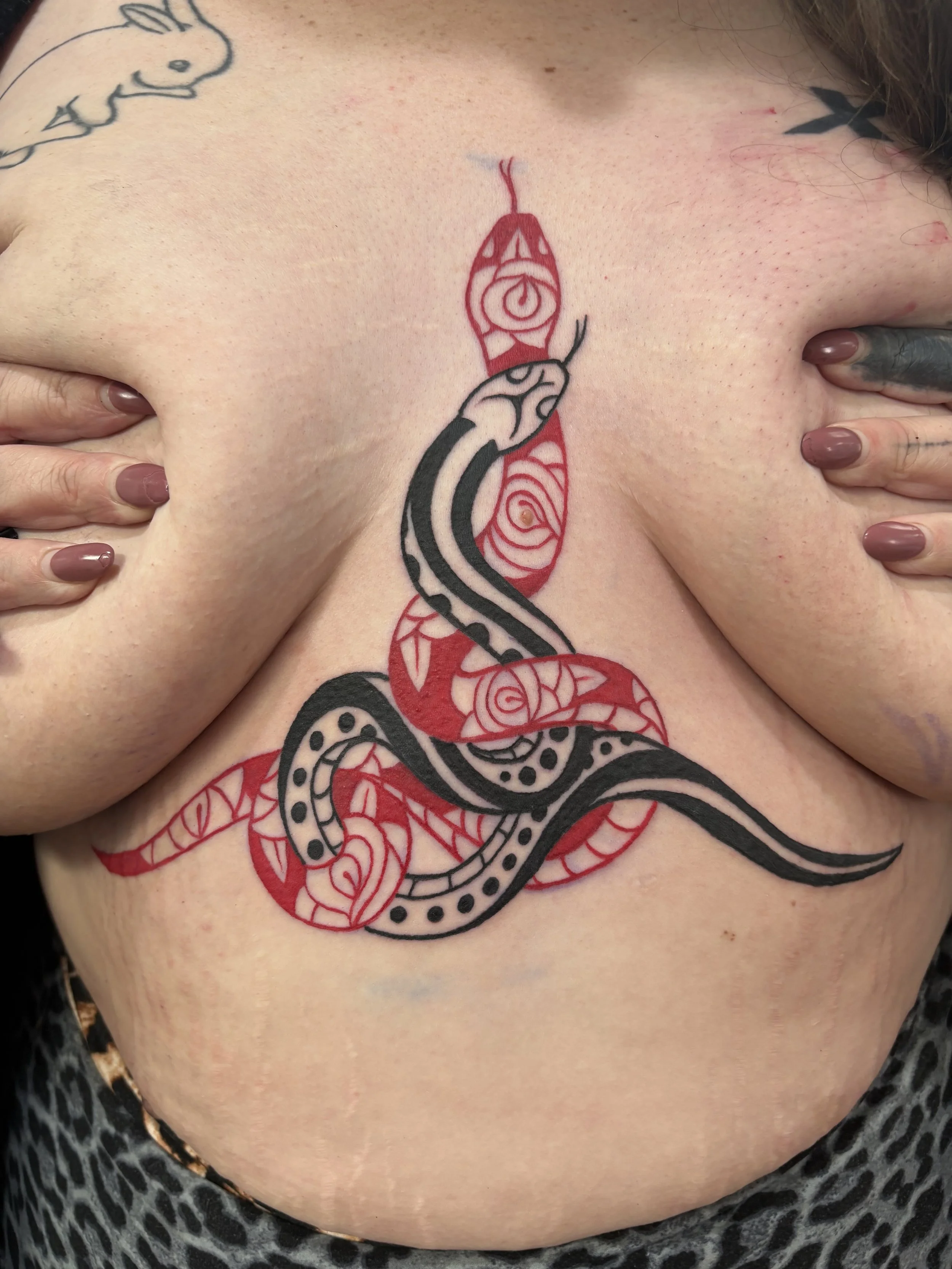 Tattoo of a stylized snake with intricate patterns in black and red ink on person's chest, with hands touching the tattoo. State Of The Art Tattooing Winchester Virginia 